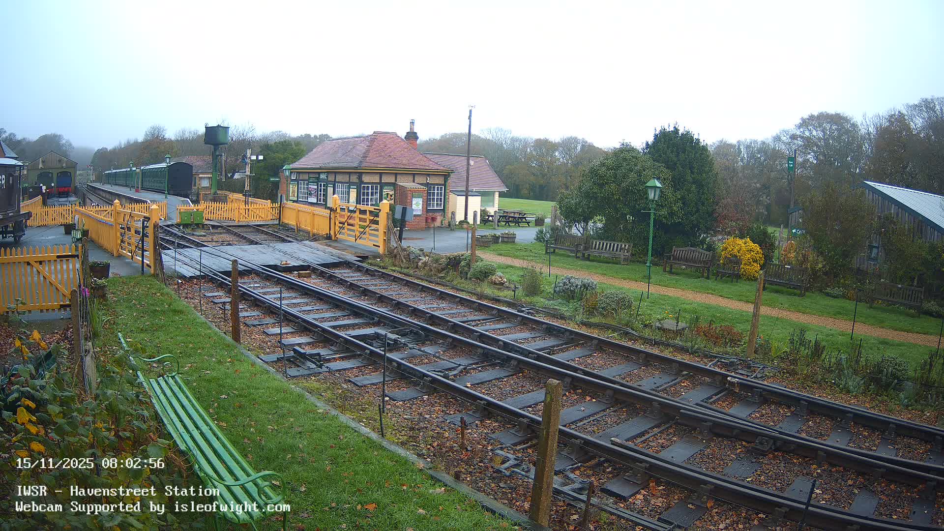 A wide view of a historic train station on an overcast autumn day, featuring multiple tracks strewn with fallen leaves in the foreground, a green train parked at a platform, a traditional station building, and wooded areas with benches in the background.