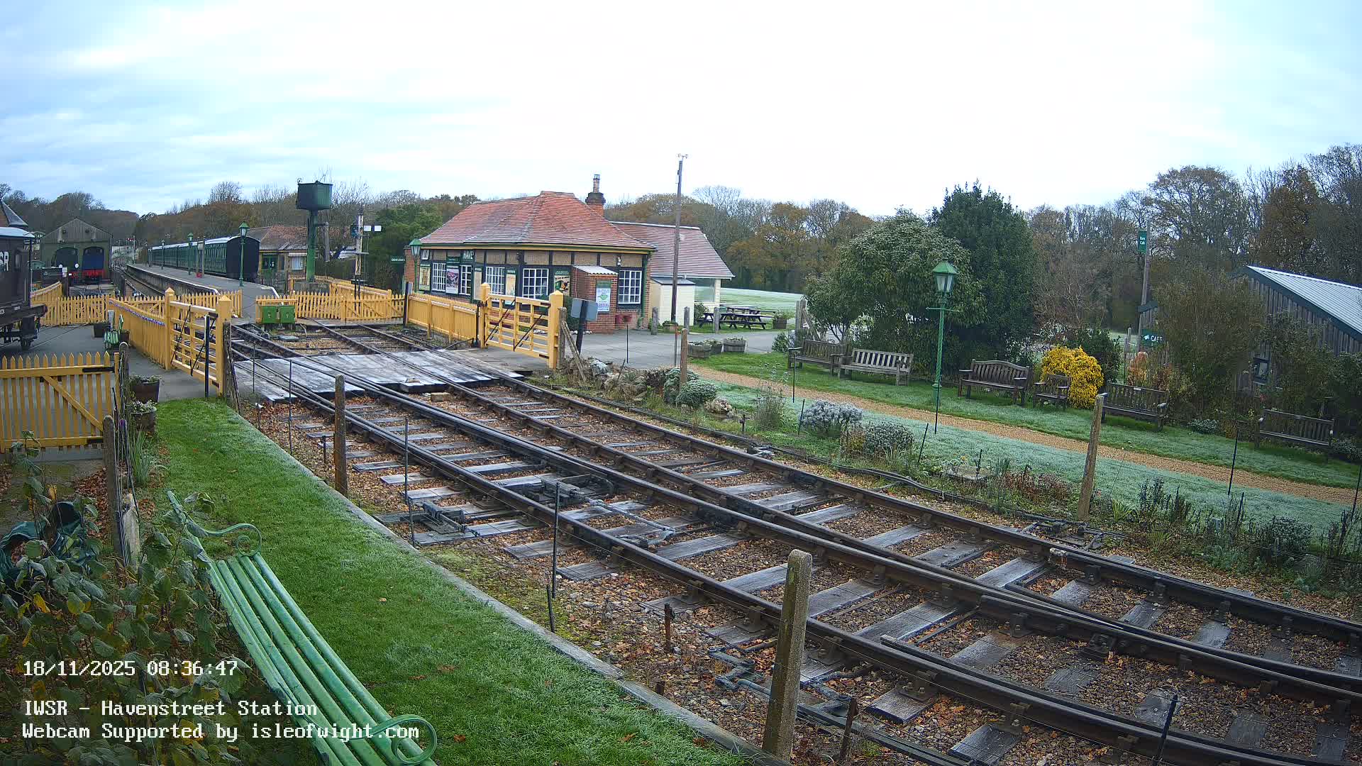A wide view of a historic train station on an overcast autumn day, featuring multiple tracks strewn with fallen leaves in the foreground, a green train parked at a platform, a traditional station building, and wooded areas with benches in the background.