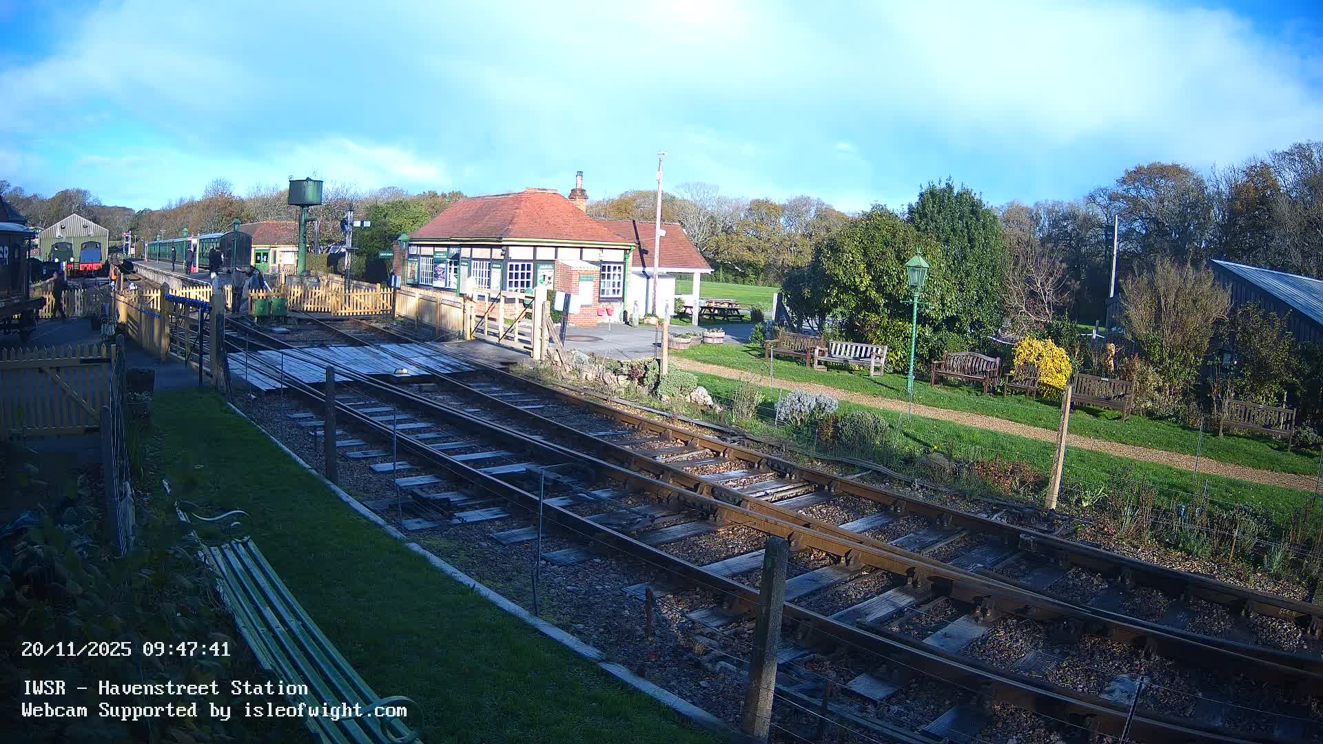On a clear, sunny day, a green train is stopped at a charming, historic station platform with passengers, multiple tracks in the foreground, and station buildings nestled among trees.
