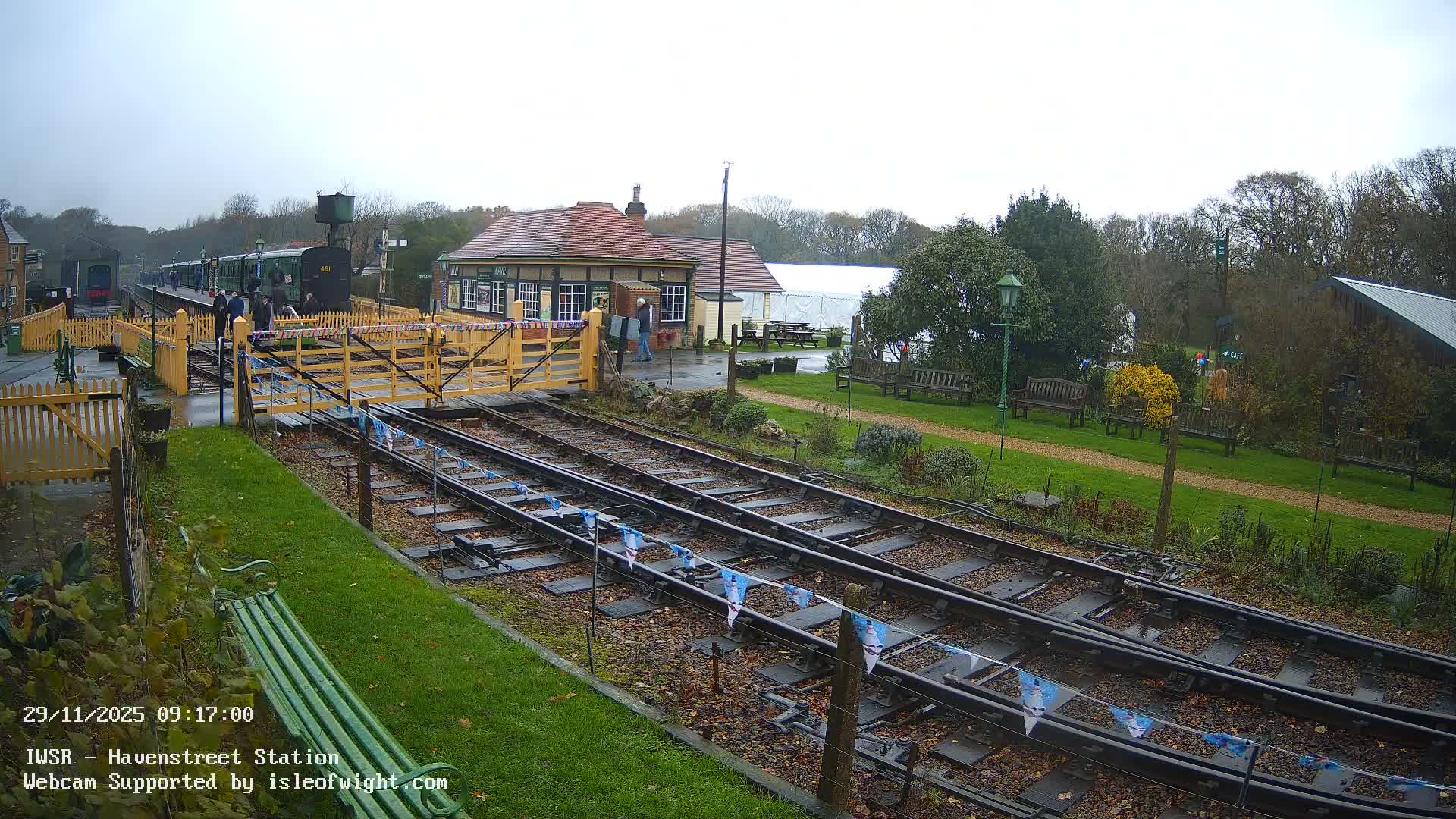 Under an overcast and damp sky, a heritage railway station features a green train at the platform with people, yellow picket fences, multiple tracks, and a traditional station building surrounded by greenery.
