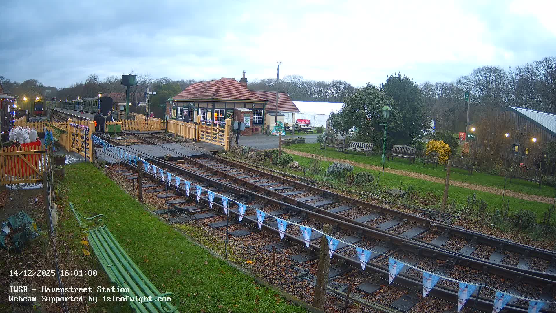 Under an overcast and damp sky, a heritage railway station features a green train at the platform with people, yellow picket fences, multiple tracks, and a traditional station building surrounded by greenery.