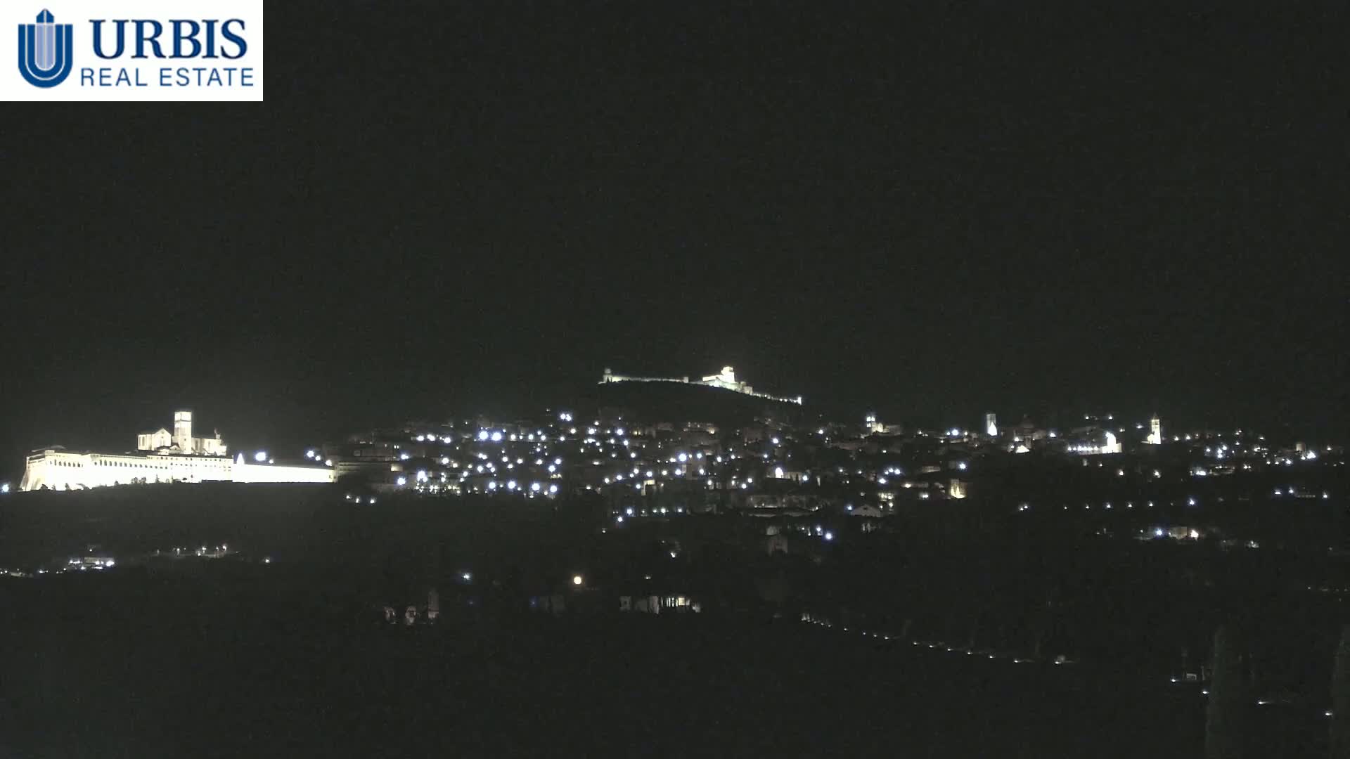 A nighttime view of a hilltop town, illuminated by many small lights, with a large, brightly lit building at the base of the hill.