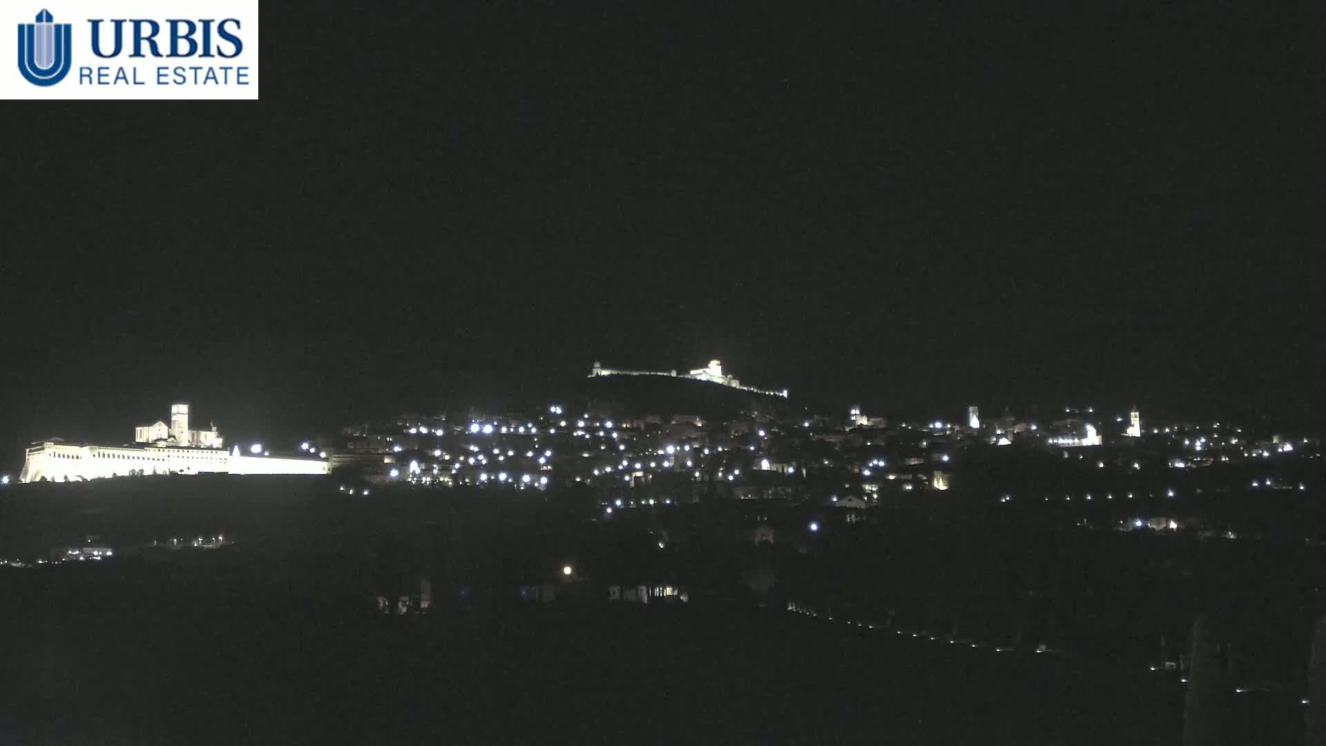 A nighttime view shows a hilltop town illuminated by scattered lights, with a large, brightly lit structure at the base of the hill.