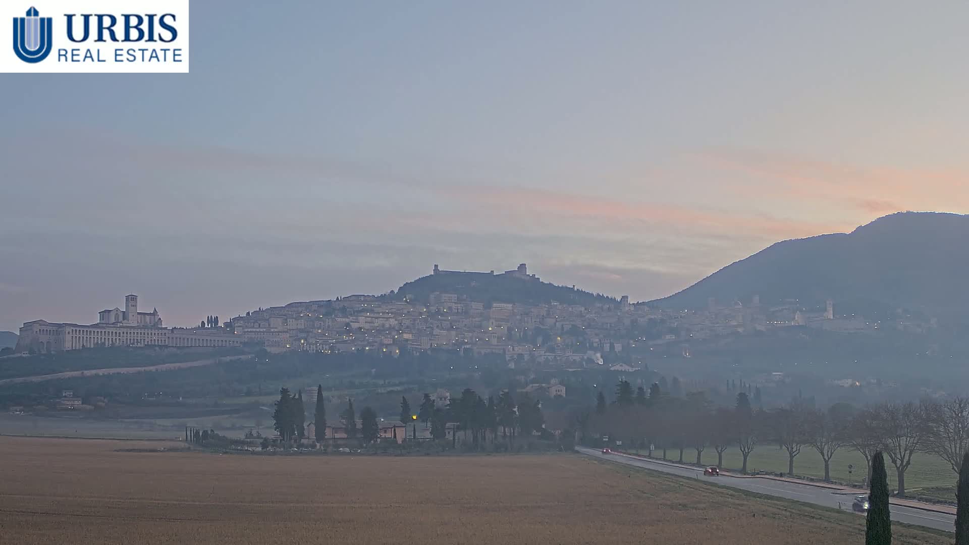 A hazy sunrise illuminates a hilltop town with a large basilica, nestled between a valley and a mountain.