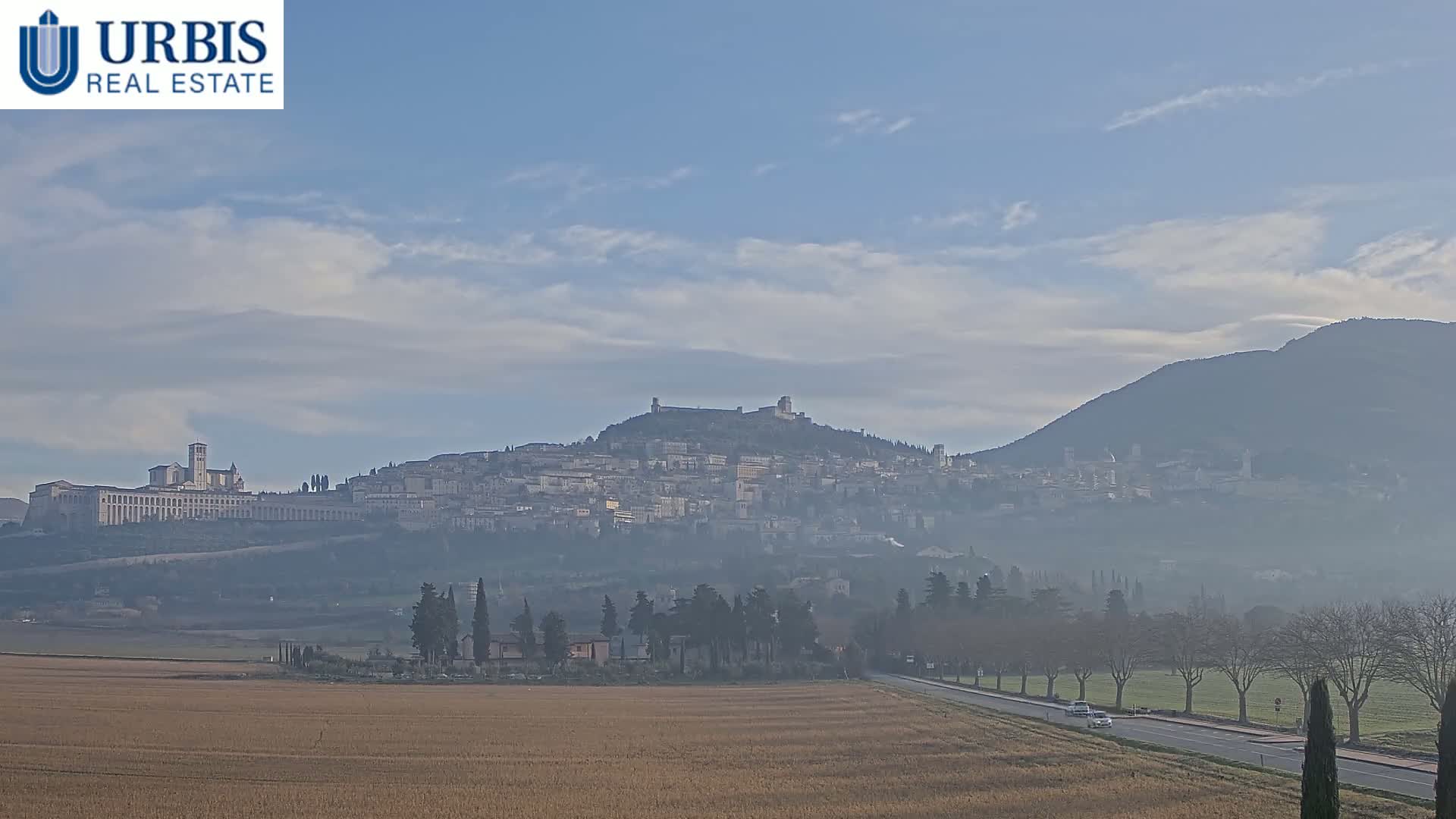 A hazy, partly cloudy day reveals a hilltop town with a large basilica, viewed from a flat, tan field with a road and trees in the foreground.