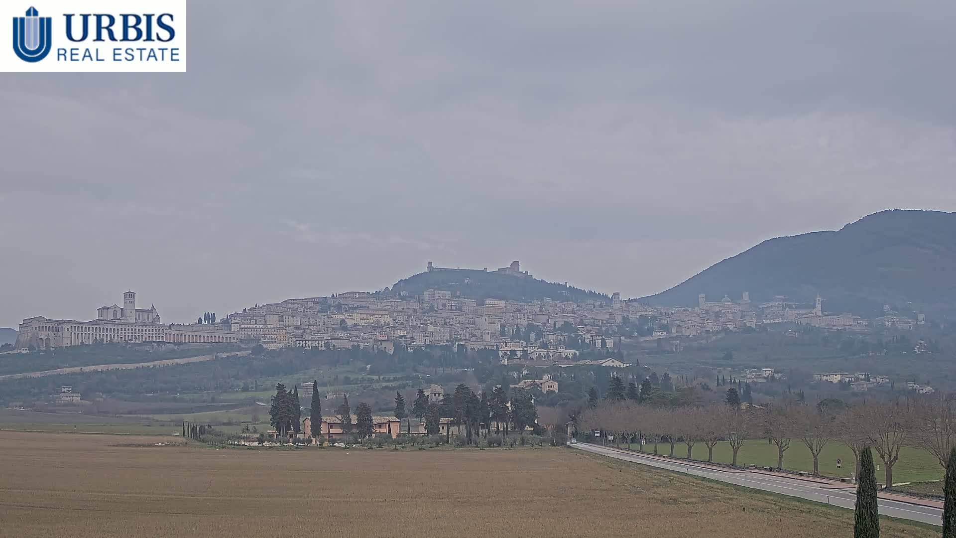 A distant view of a hilltop town, under a hazy, overcast sky, stretches across a flat, brown field with a road leading towards it.