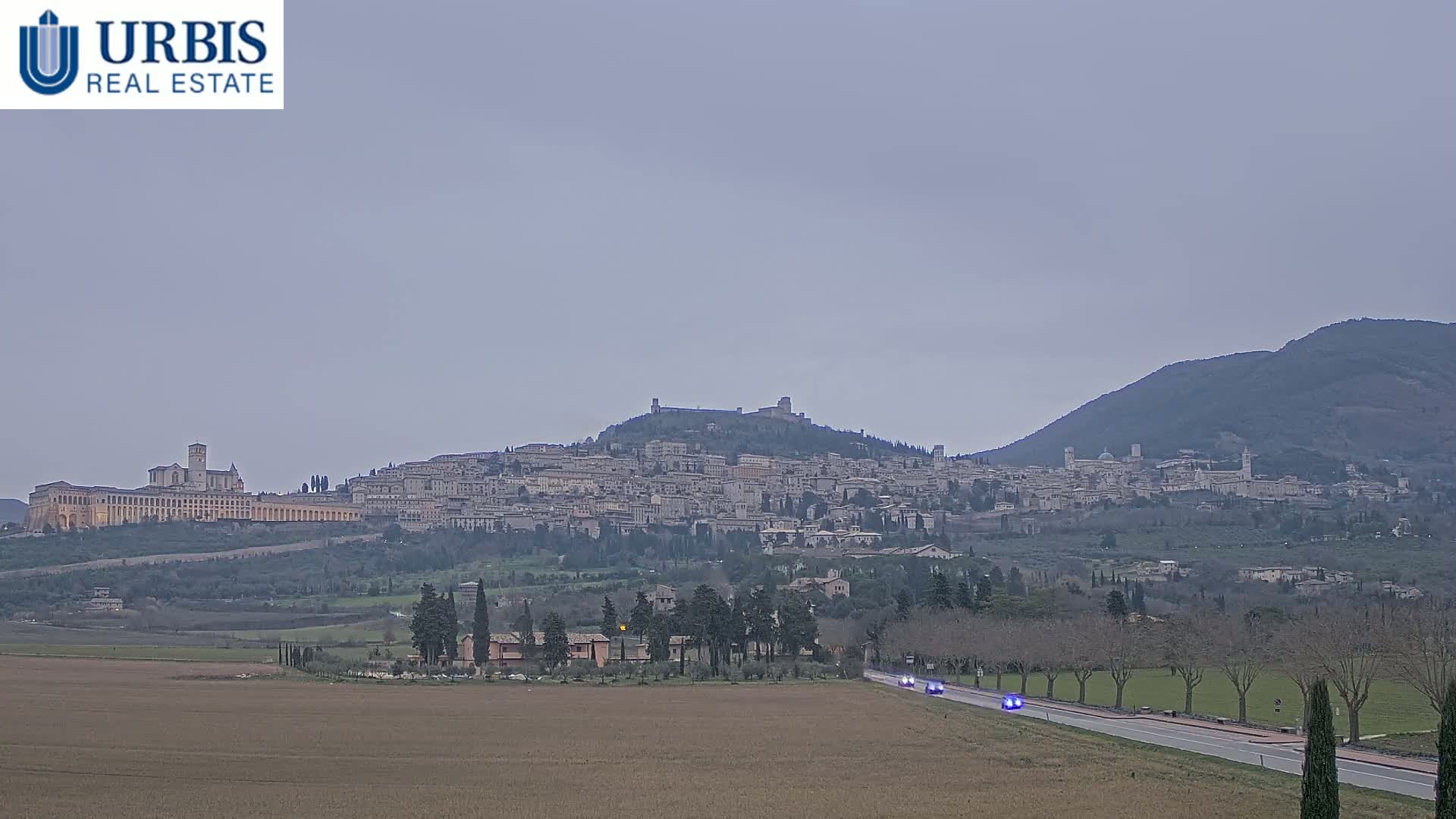 A hilltop town, possibly Assisi, is visible in the distance under an overcast sky, with a road and sparse vegetation in the foreground.