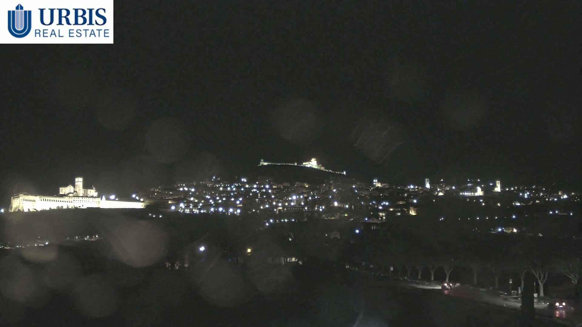 A nighttime view of a hilltop town illuminated with lights, seen through rain-covered glass.