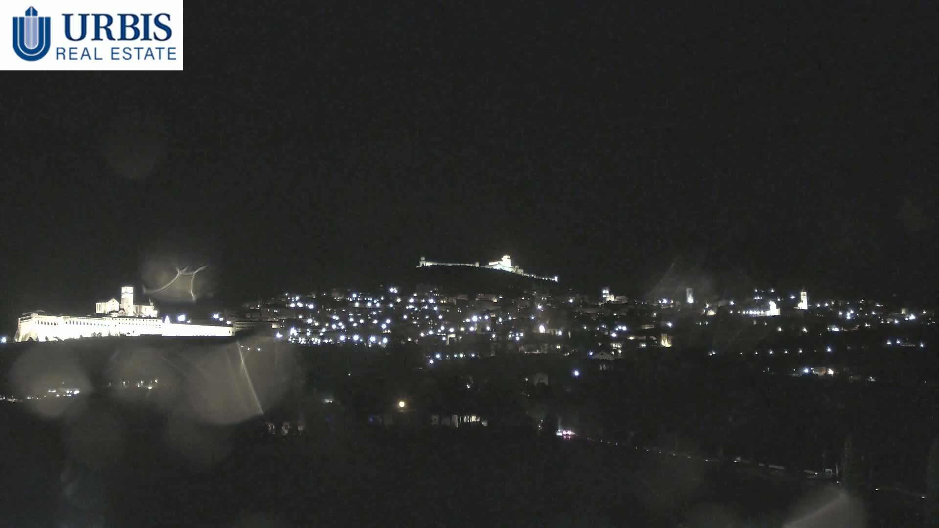 A nighttime view of a hilltop town illuminated by scattered lights, with rain obscuring the foreground.