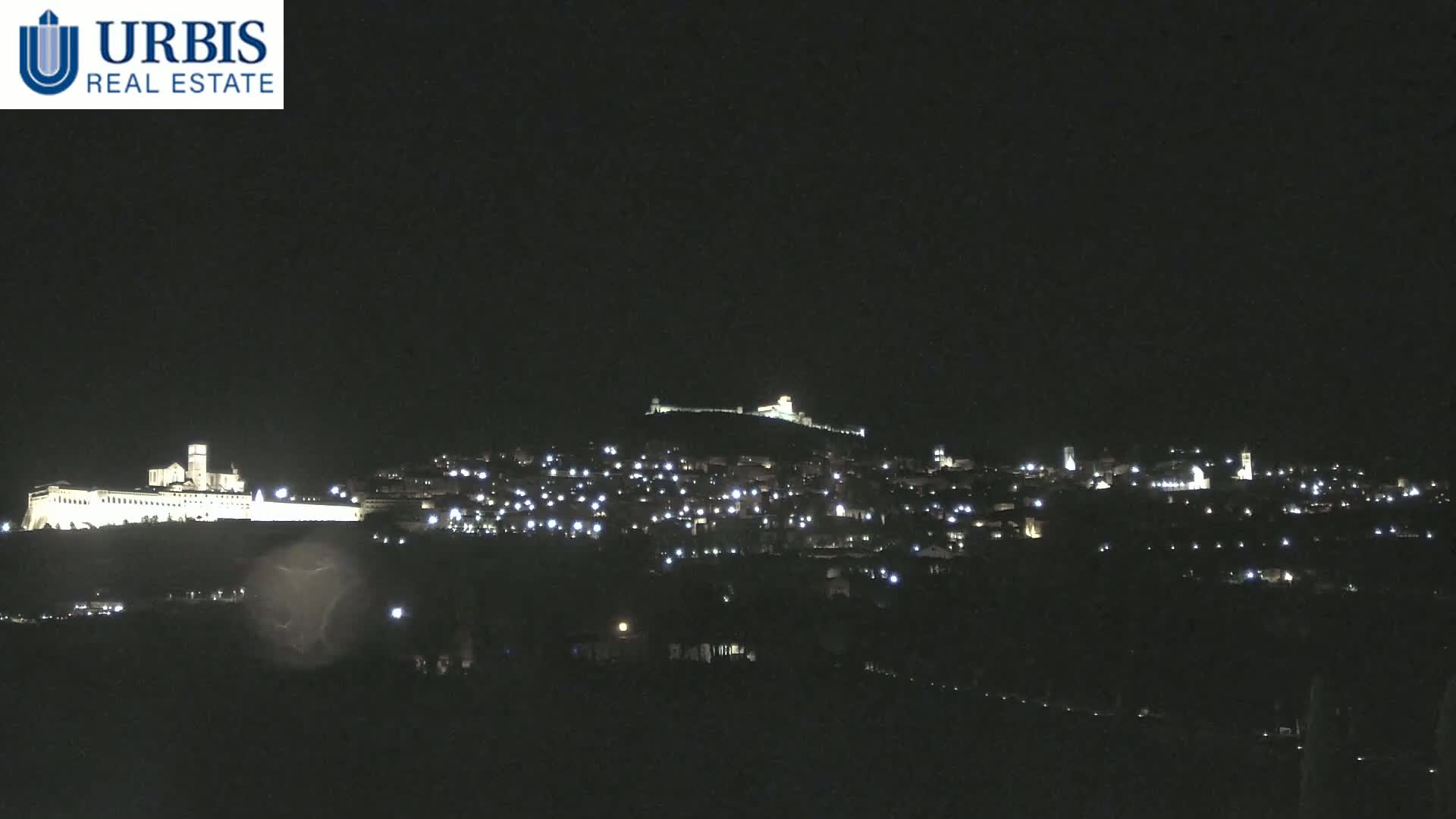 A hilltop town is illuminated at night, with a large, brightly lit structure on a lower hill to the left.