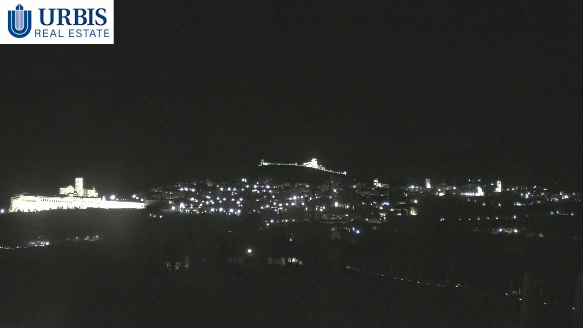A nighttime view of a hilltop town illuminated by lights, with a large, brightly lit building on a lower hill in the foreground.