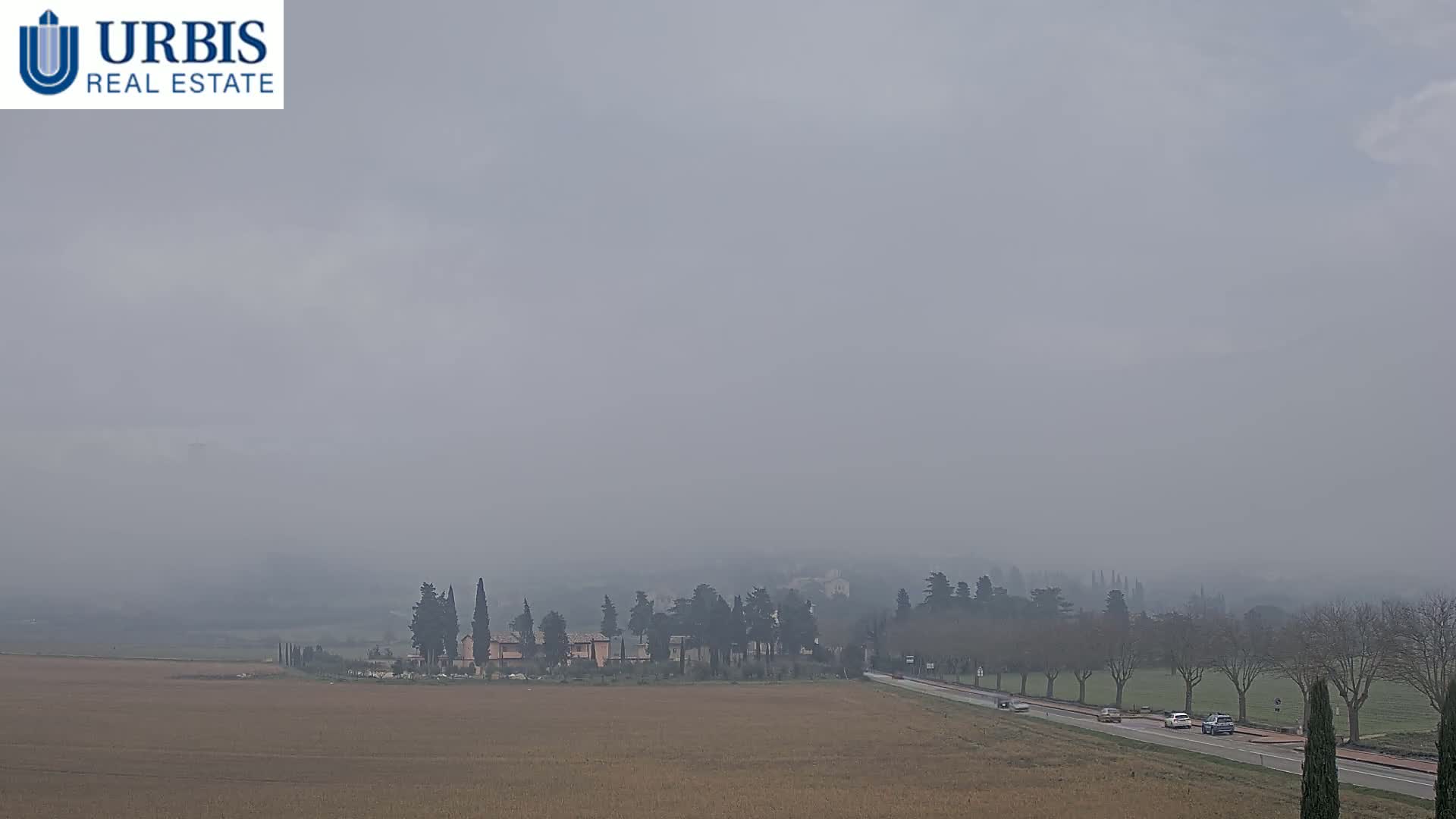 A hazy, overcast day reveals a flat, brown field, a road with a few cars, and a small cluster of buildings surrounded by trees in the distance.