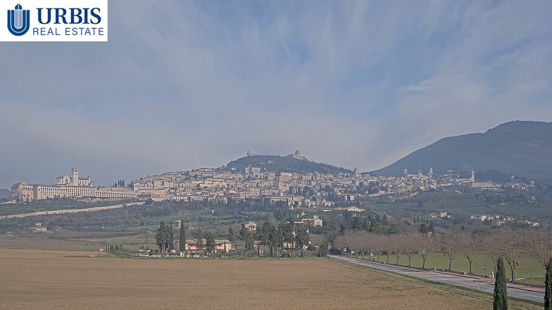 A hilltop town with many buildings, including a large basilica, is visible in the distance under a mostly clear sky, with a flat field and a road in the foreground.