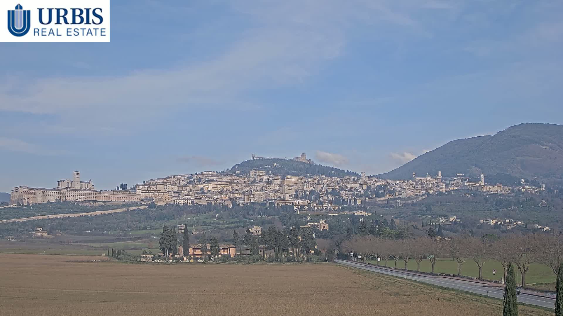 A large hilltop town, under a mostly clear blue sky, is visible from a distance across a flat field and a road.