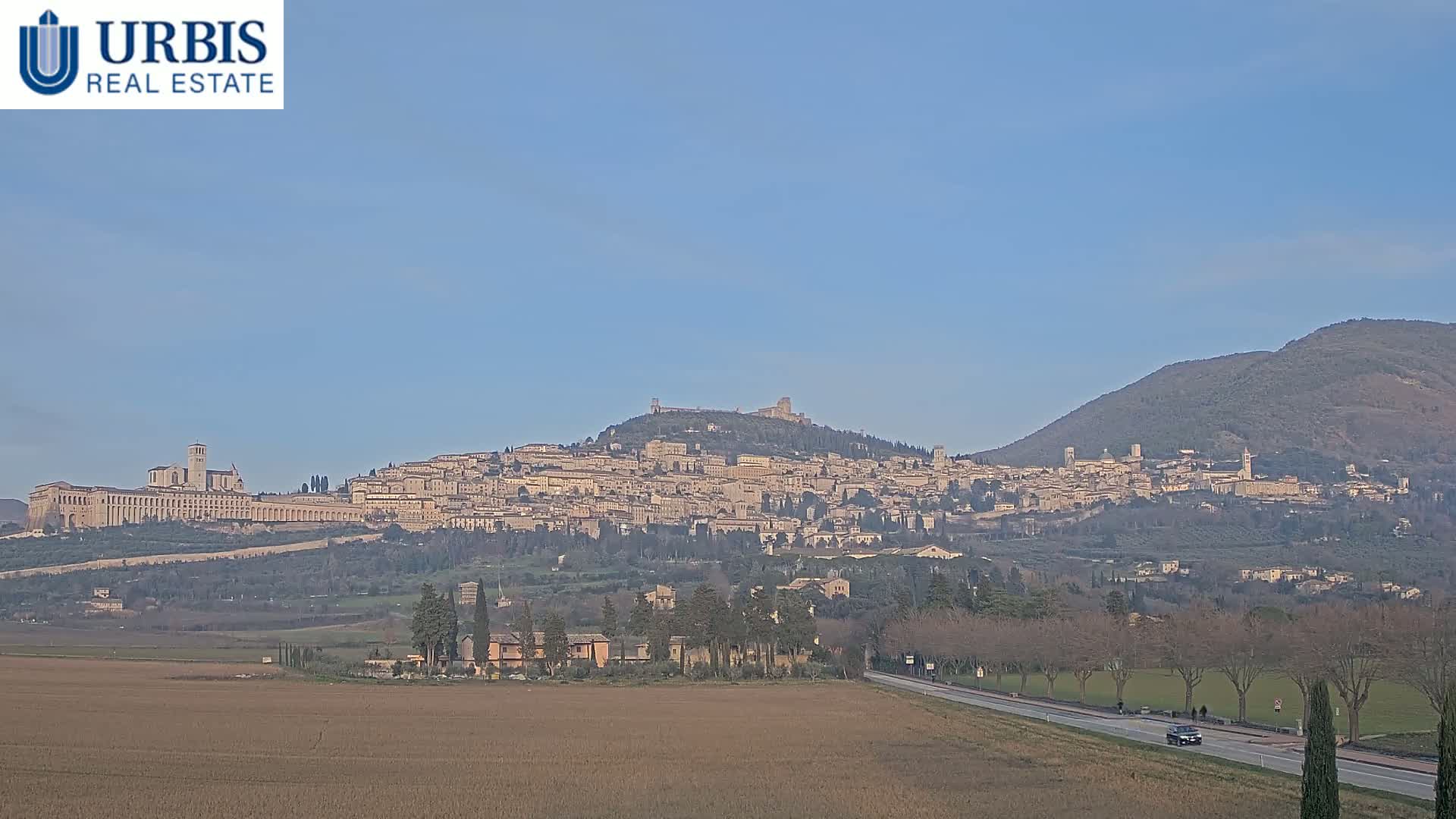 A large hilltop town, possibly Assisi, is visible in the distance under a clear blue sky, with a flat, brown field in the foreground.