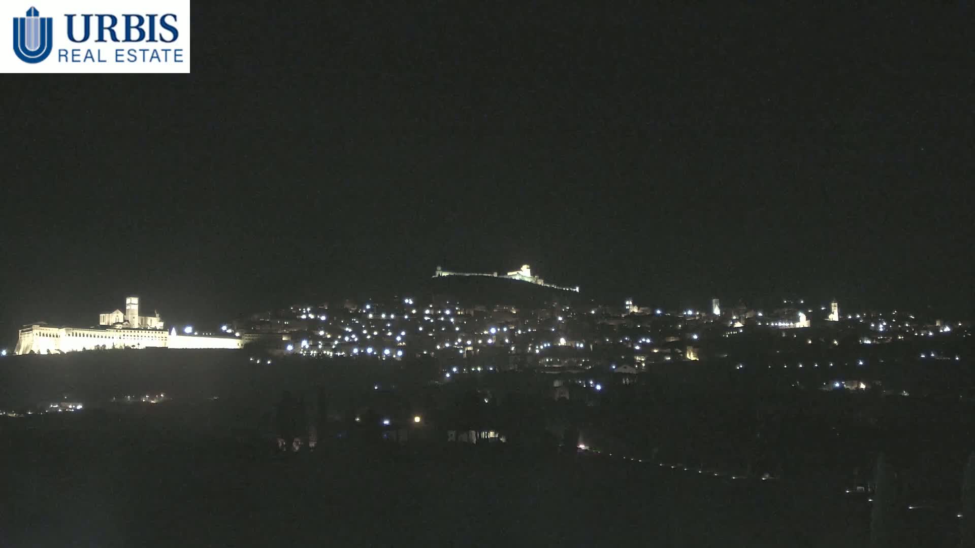 A nighttime view of a hilltop town illuminated by scattered lights, with a large, brightly lit building complex on a lower hill in the background.
