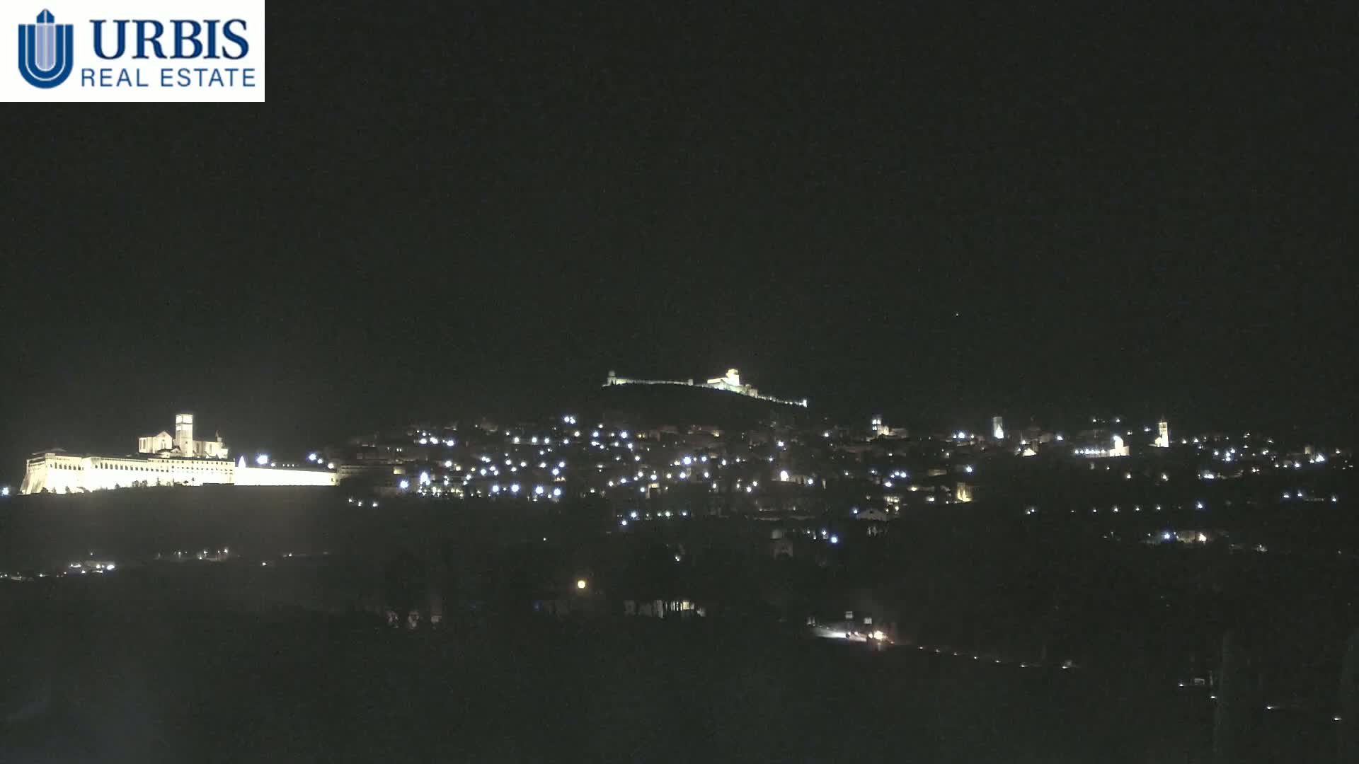 A nighttime long shot shows a hilltop town illuminated with scattered lights, including a large, brightly lit building on a lower hill in the foreground.