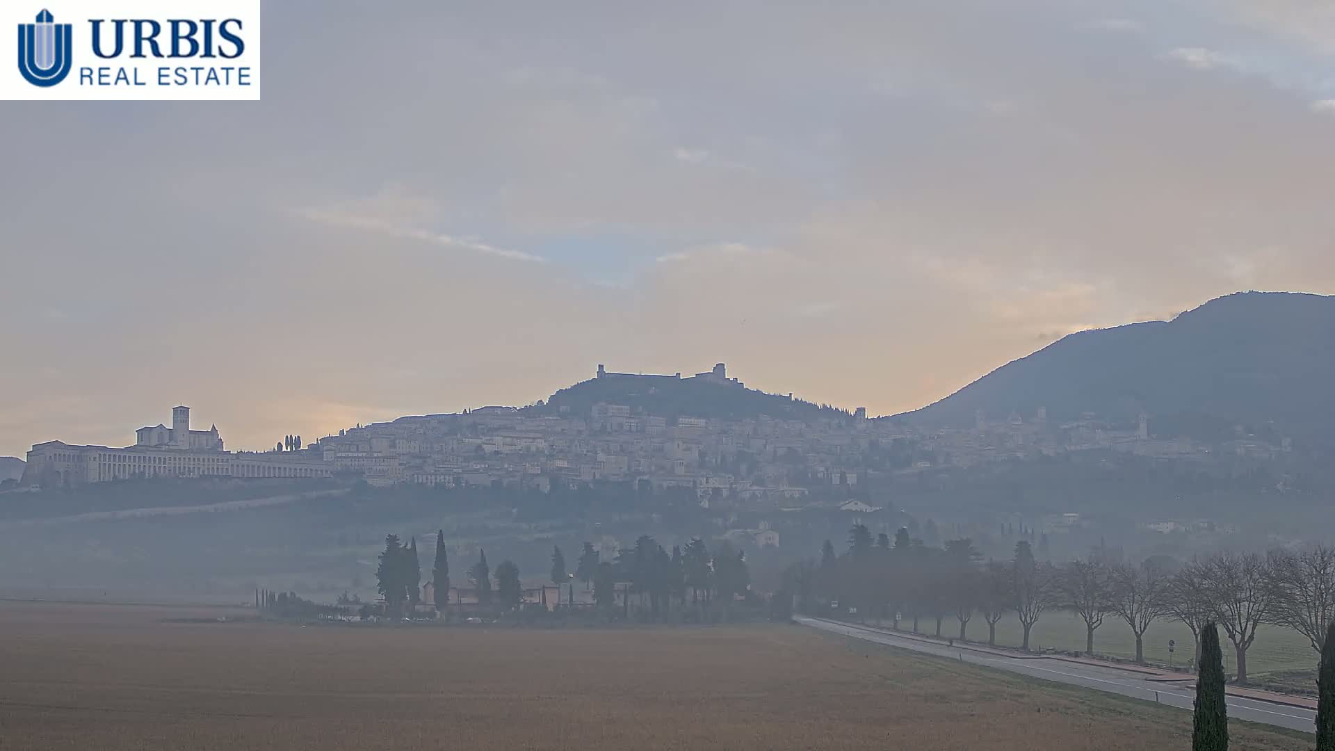 A hazy, distant view of a hilltop town, partially obscured by fog, with a large, flat-topped structure prominent in the foreground.