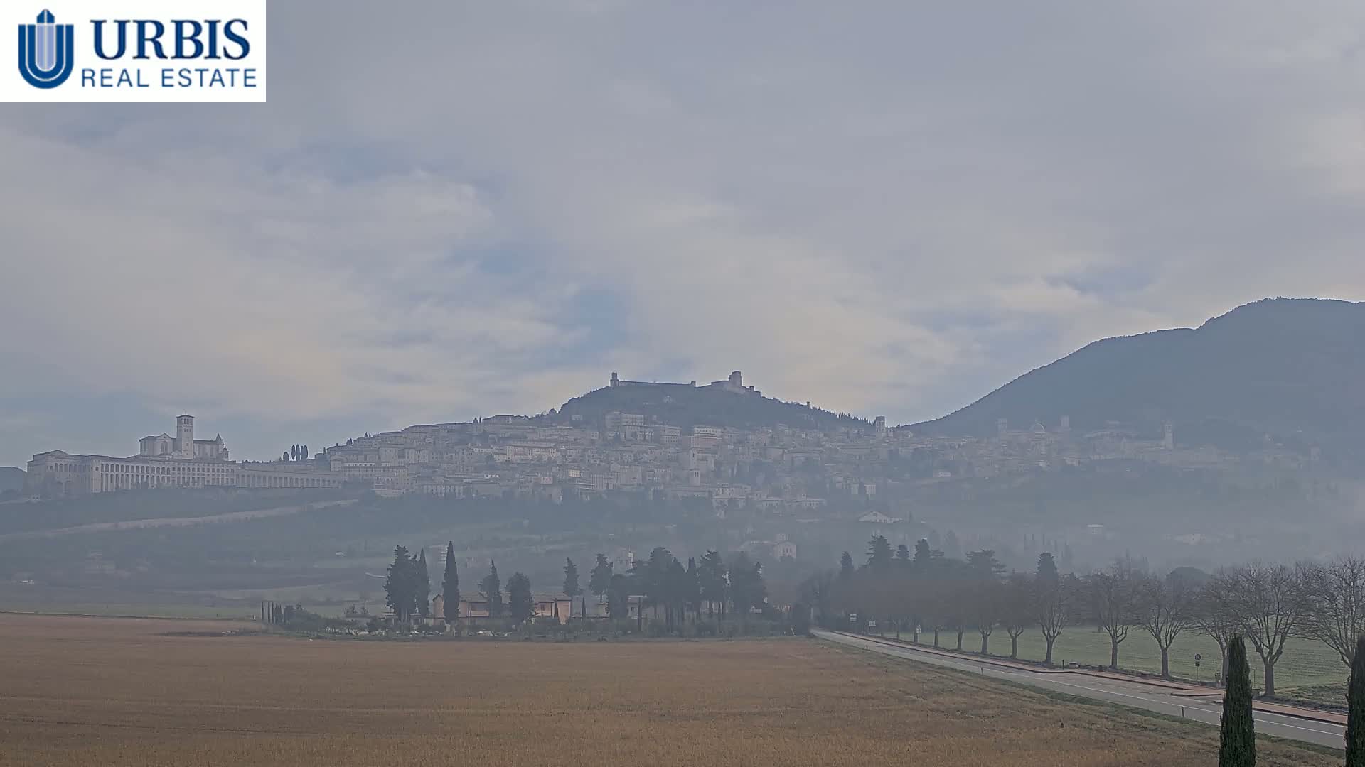 A hazy, overcast day reveals a hilltop town nestled between two larger hills, with a flat field and road in the foreground.