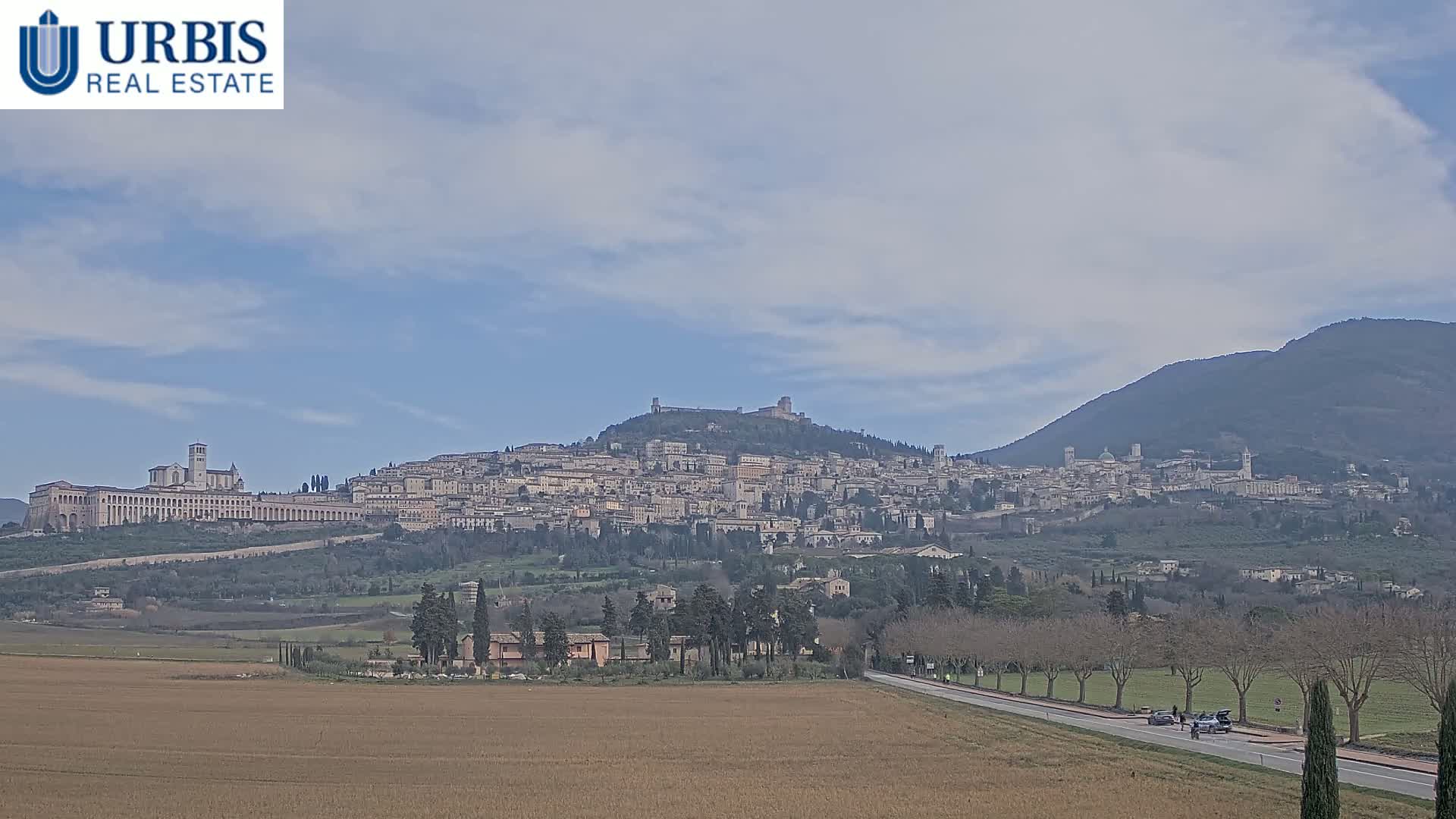 A hilltop town, nestled between a flat field and a mountain, under a mostly clear, light-blue sky.