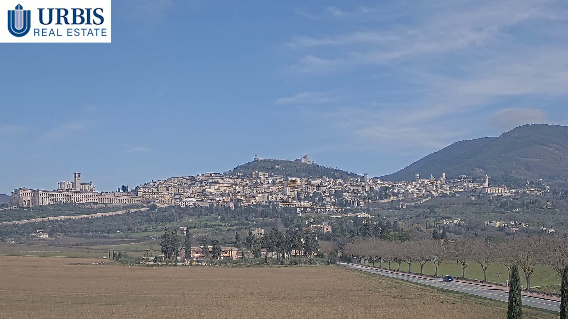 A sprawling hilltop town, under a mostly clear blue sky, is viewed from a distance across a flat, brown field and a roadway.