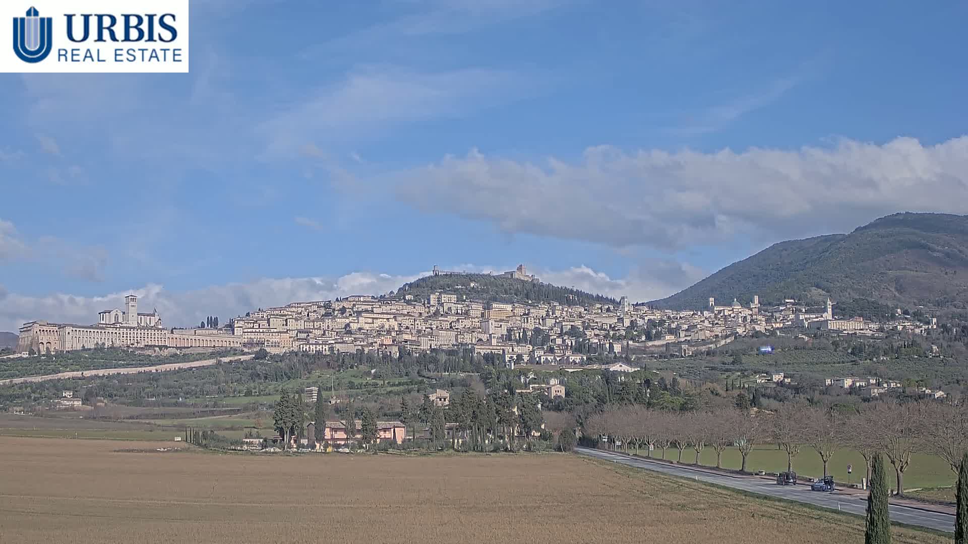 A sprawling hilltop town, under a partly cloudy sky, is seen from a distance across a flat, brown field and a road with a few cars.