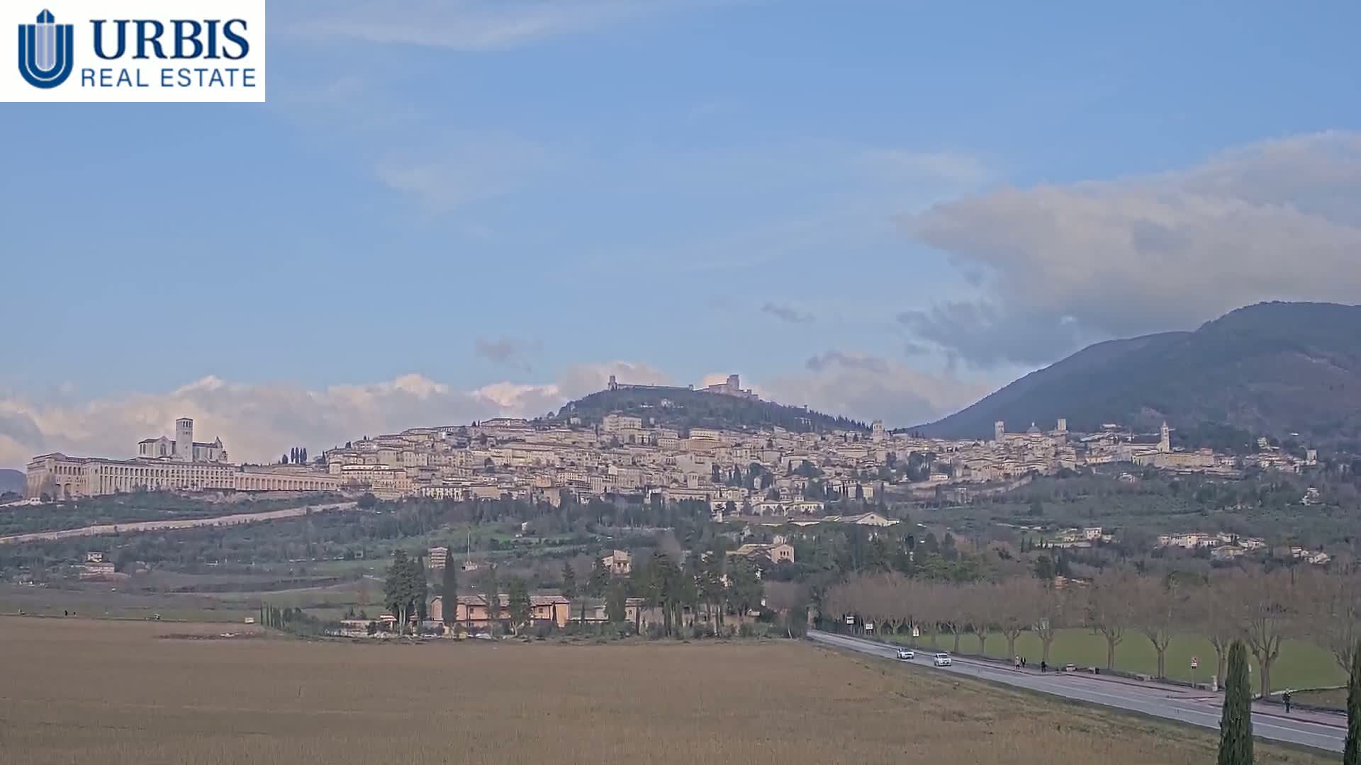 A sprawling town nestled on a hill, under a partly cloudy sky, is viewed from a distance across a flat field and a road.
