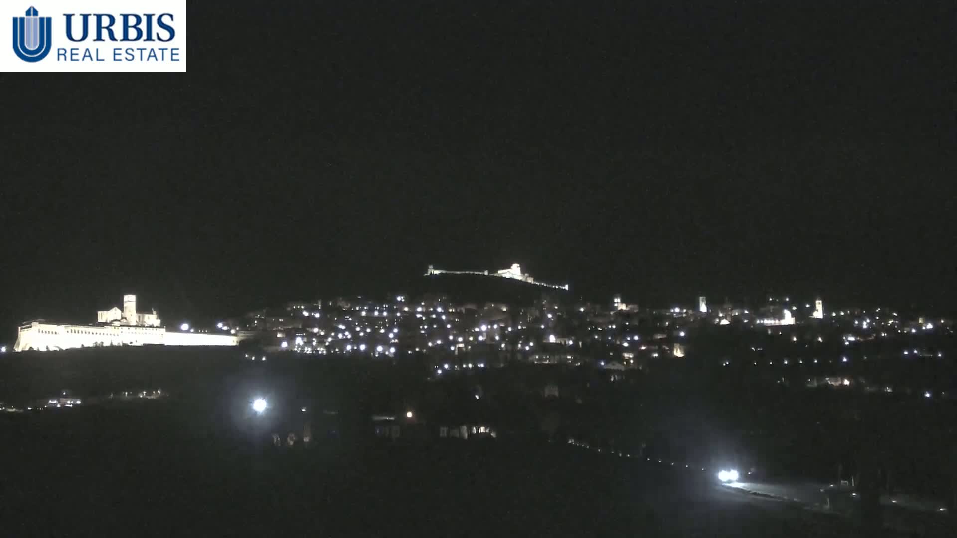 A nighttime view of a city and hilltop buildings illuminated by lights under a clear, dark sky.