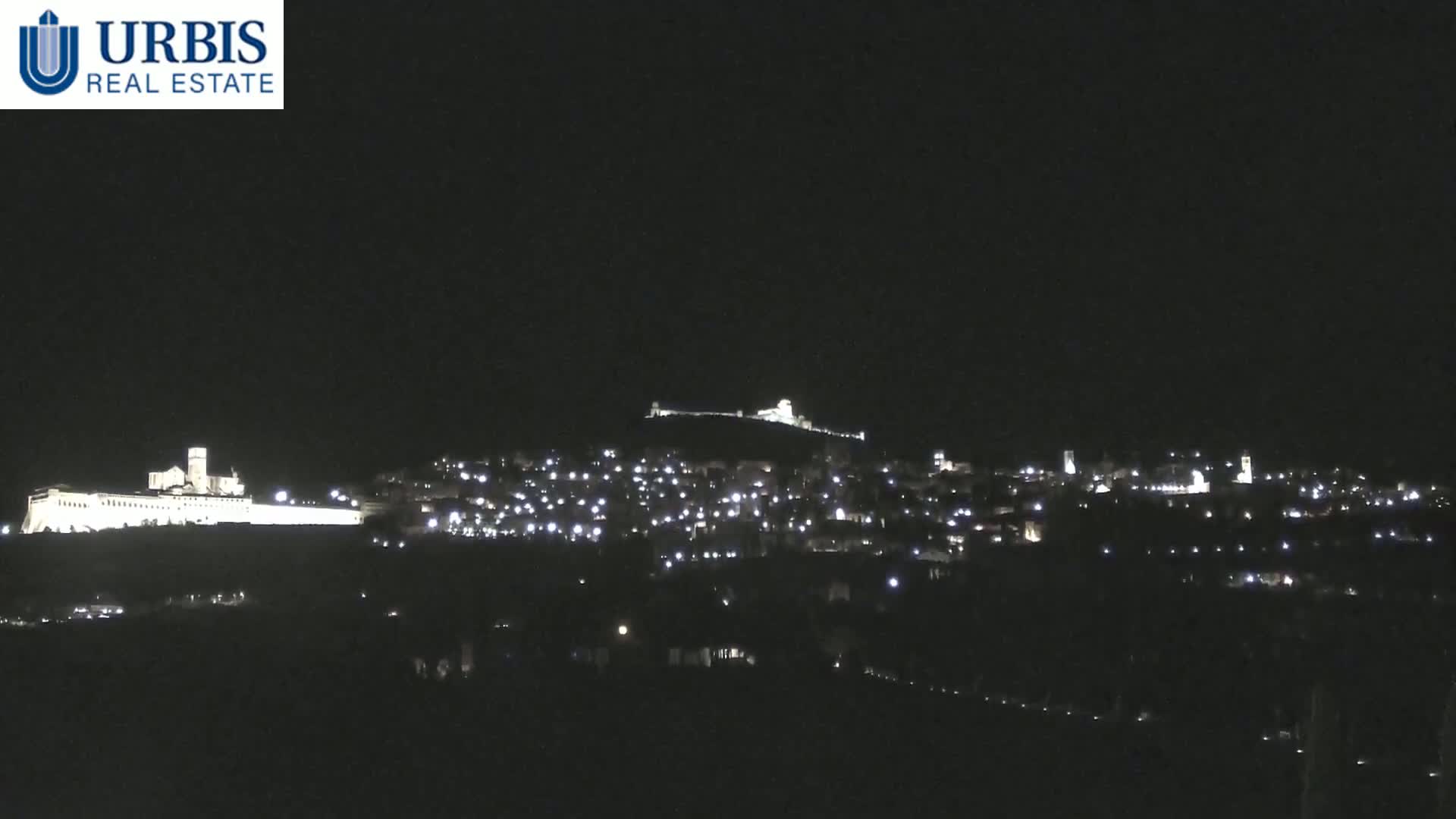 A nighttime long shot shows a hilltop town with numerous lights illuminating buildings, including a large, brightly lit structure on a lower hill.
