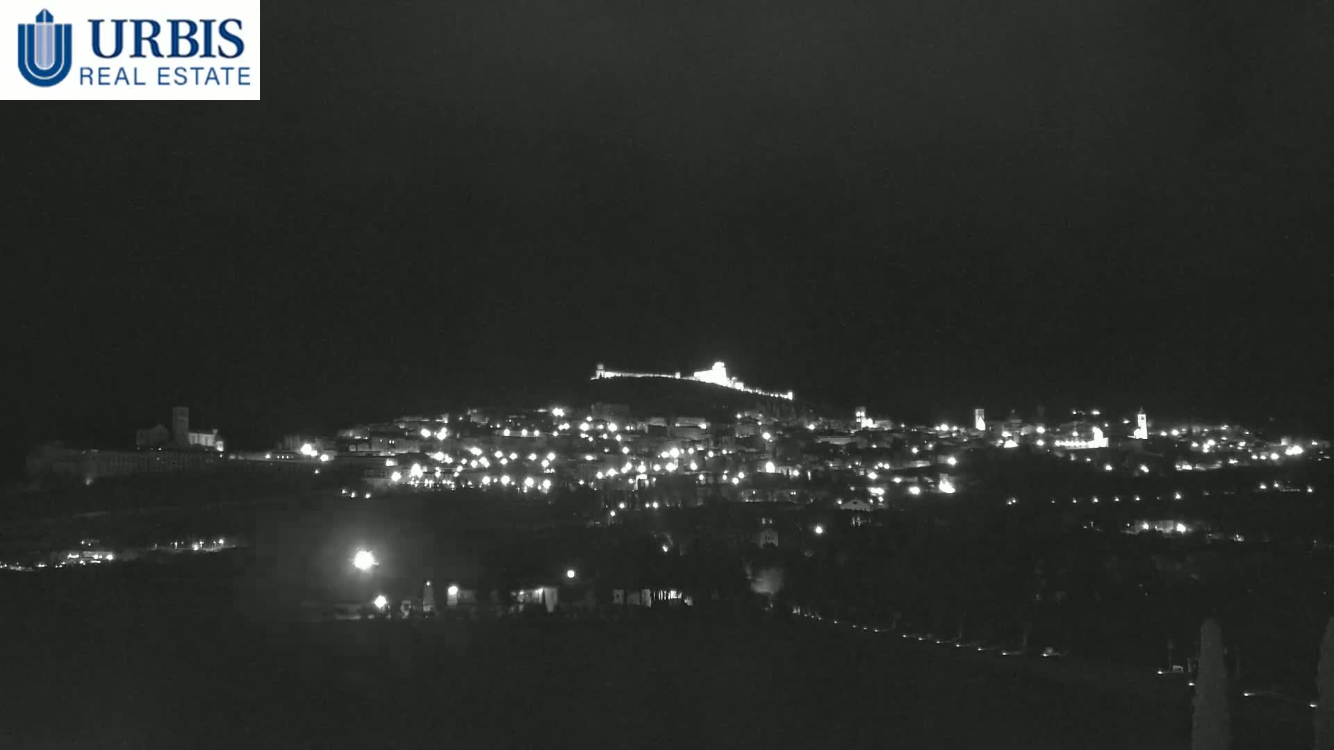 A nighttime long shot shows a hilltop town illuminated by many small lights, under a dark, clear sky.