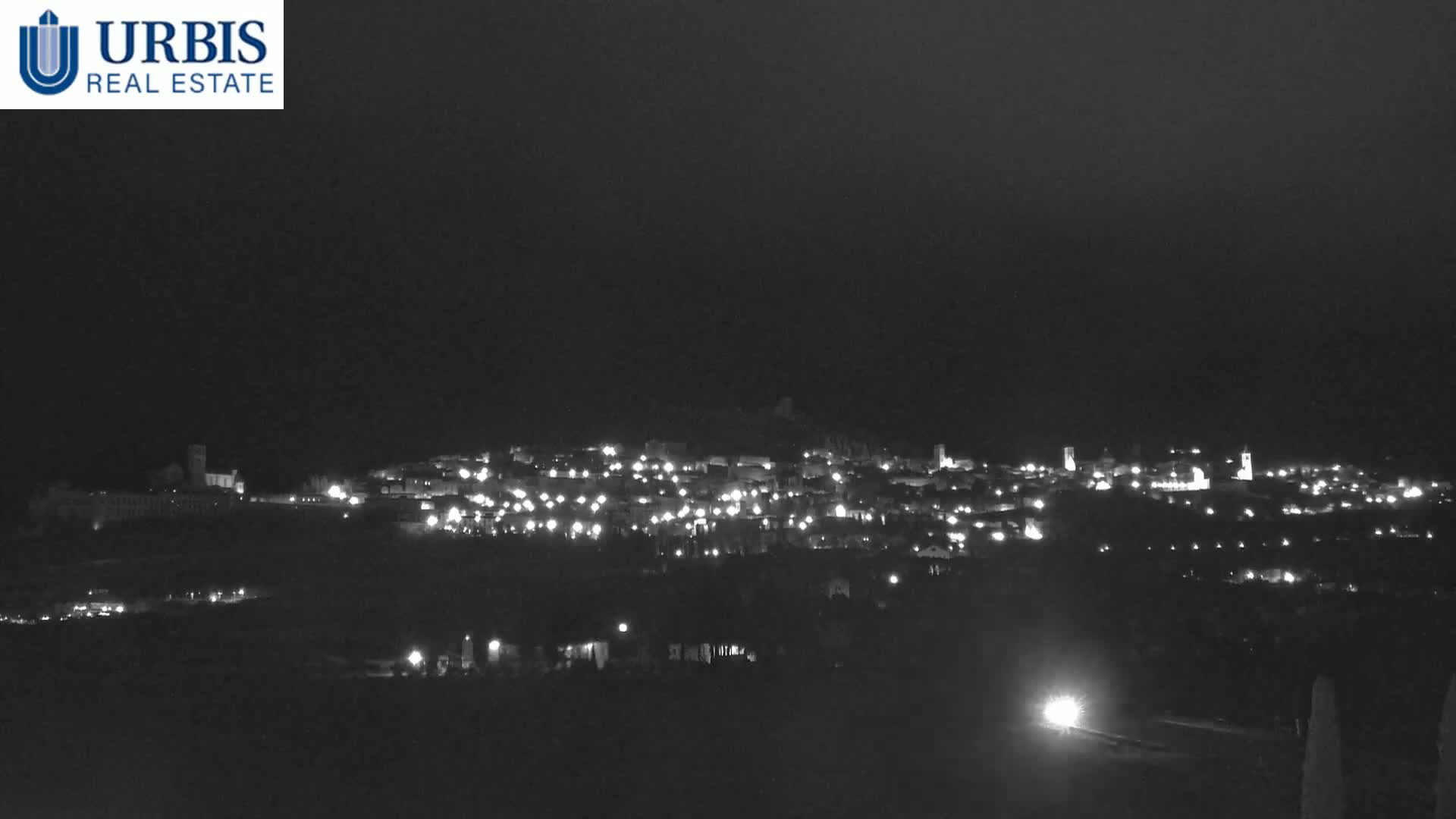 A nighttime long shot shows a town illuminated by many small lights on a dark hill under a clear, dark sky.
