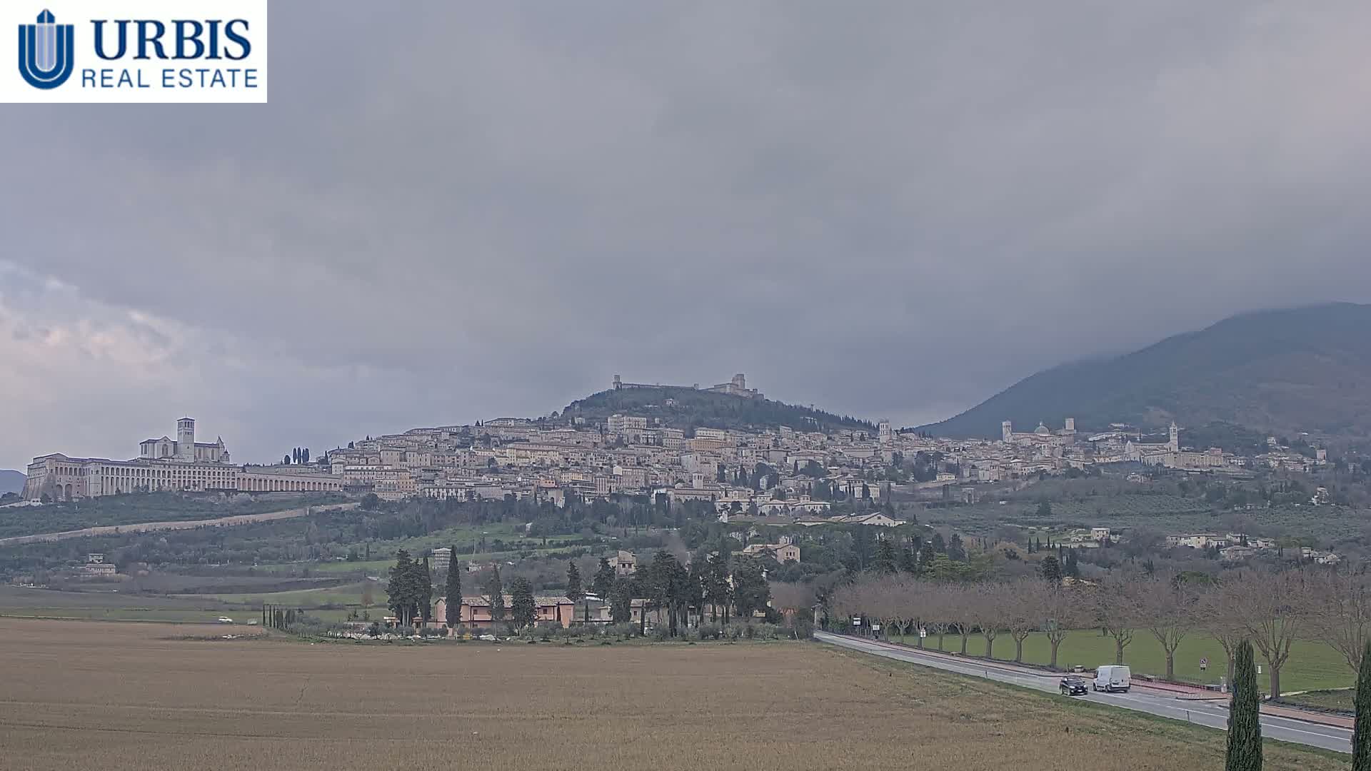 A large hilltop town, under a cloudy sky, is visible from a distance across a flat field and a road with sparse traffic.