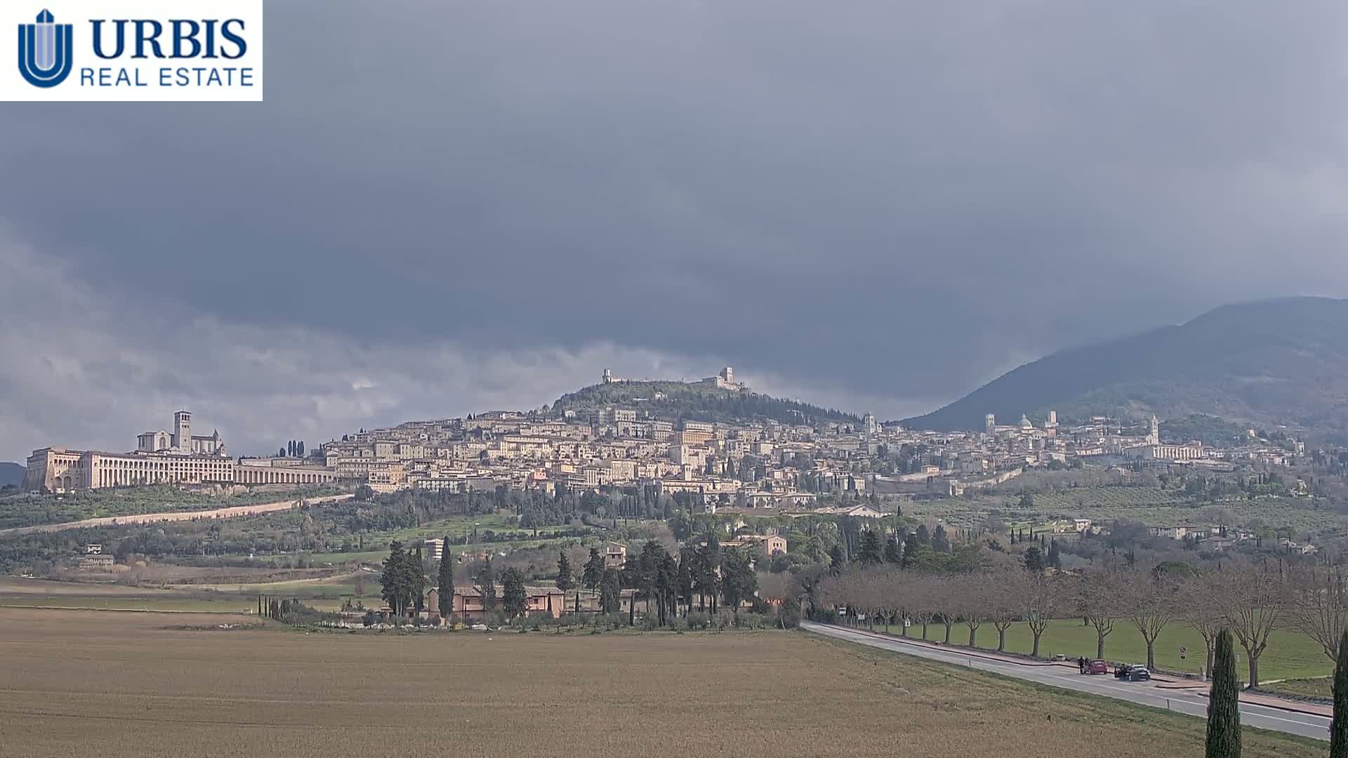 A large hilltop town is visible under an overcast sky, with a road and fields in the foreground.