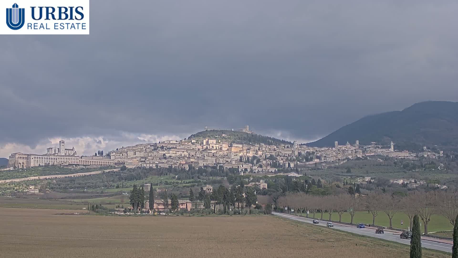 A sprawling town nestled on a hill, under a heavily clouded sky, is viewed from a distance across a flat, brown field and a road with sparse traffic.