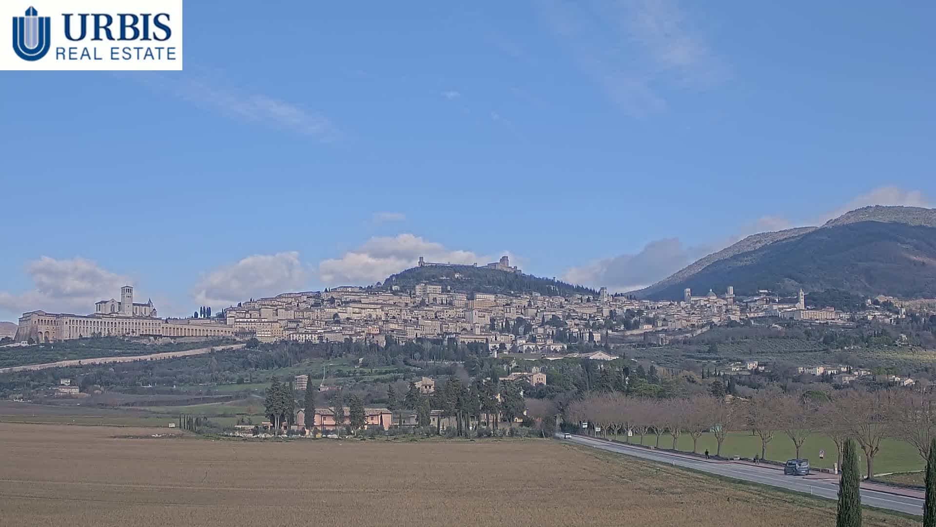 A hilltop town with numerous buildings, including a large basilica, is visible under a mostly clear blue sky, with a few clouds present.