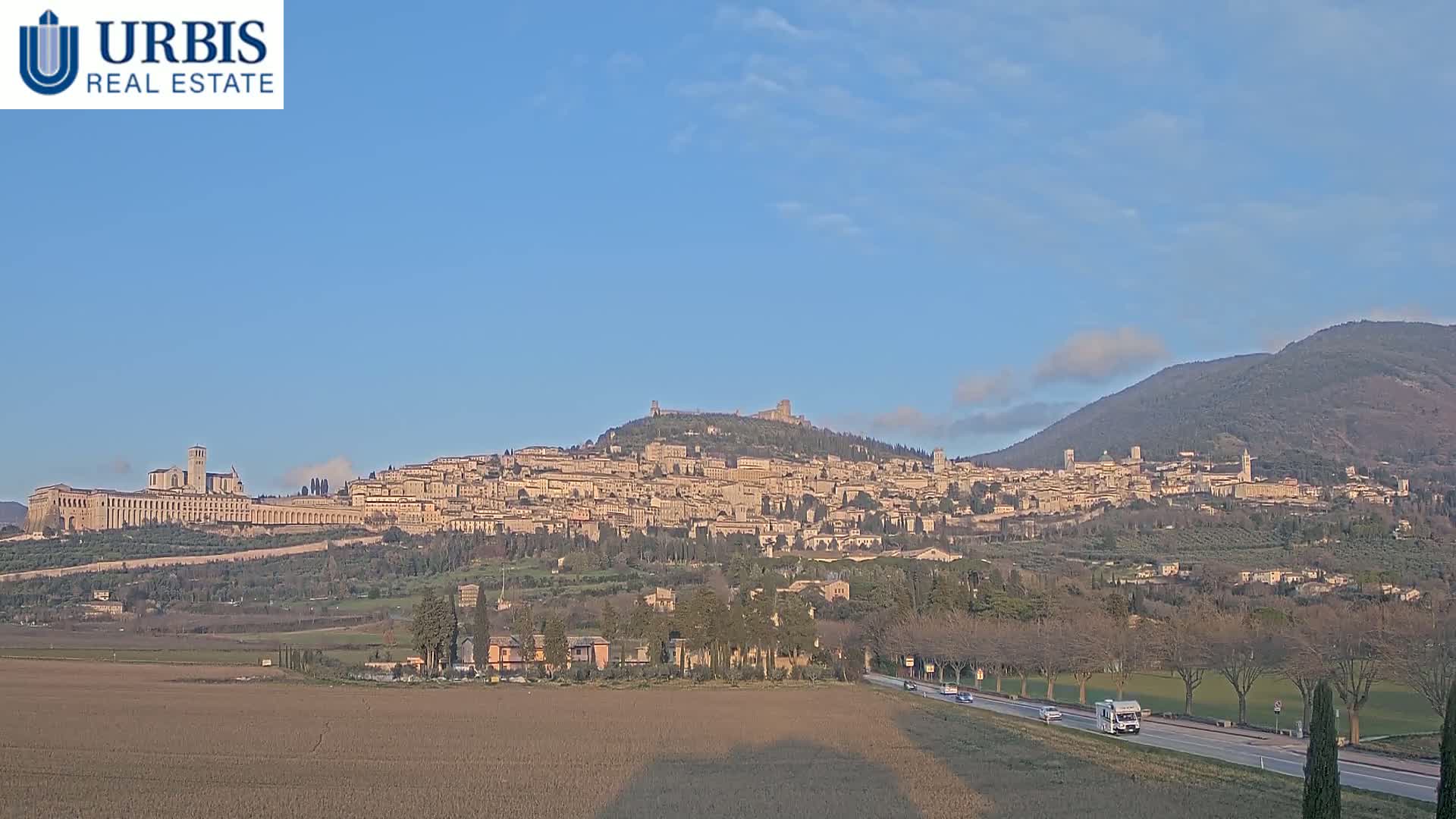 A hilltop town with numerous buildings, including a large basilica, is seen under a clear, bright sky, with a road and flatlands in the foreground and mountains in the background.
