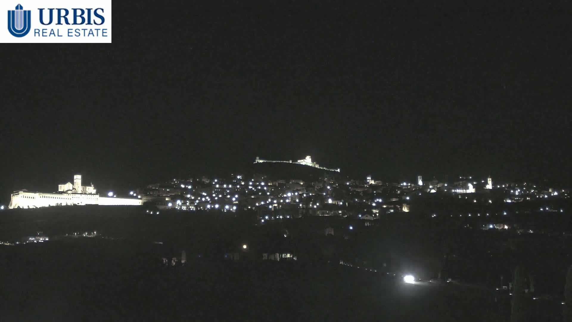 A nighttime view of a hilltop town illuminated by lights, with a large, brightly lit building on a lower hill in the foreground under a clear, dark sky.