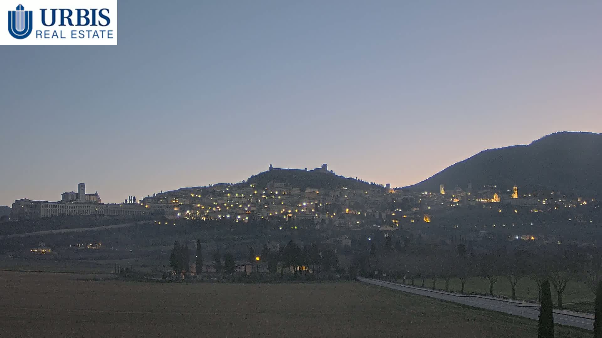 A hilltop town, illuminated at twilight, is nestled between two larger hills under a dusky sky.