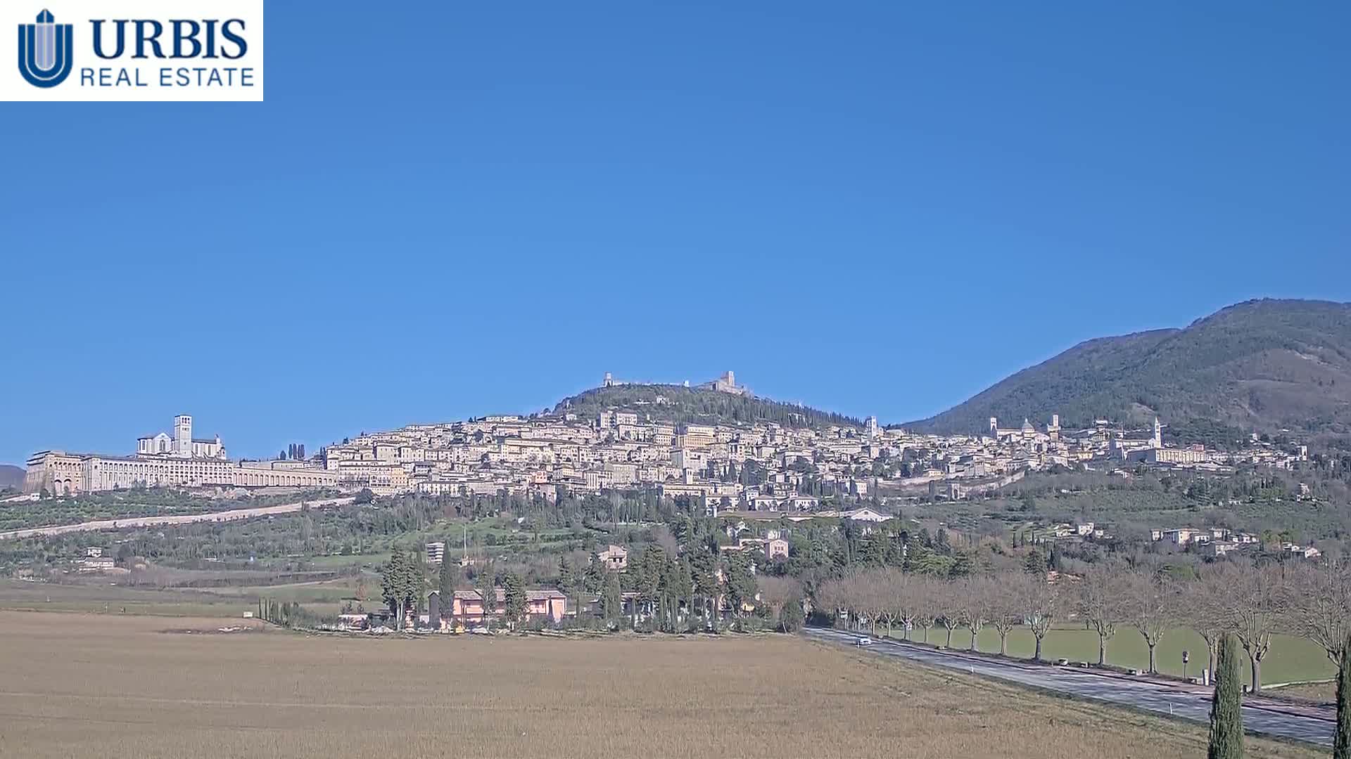 A hilltop town, nestled between mountains under a clear blue sky, is viewed from a distance across a flat, brown field.