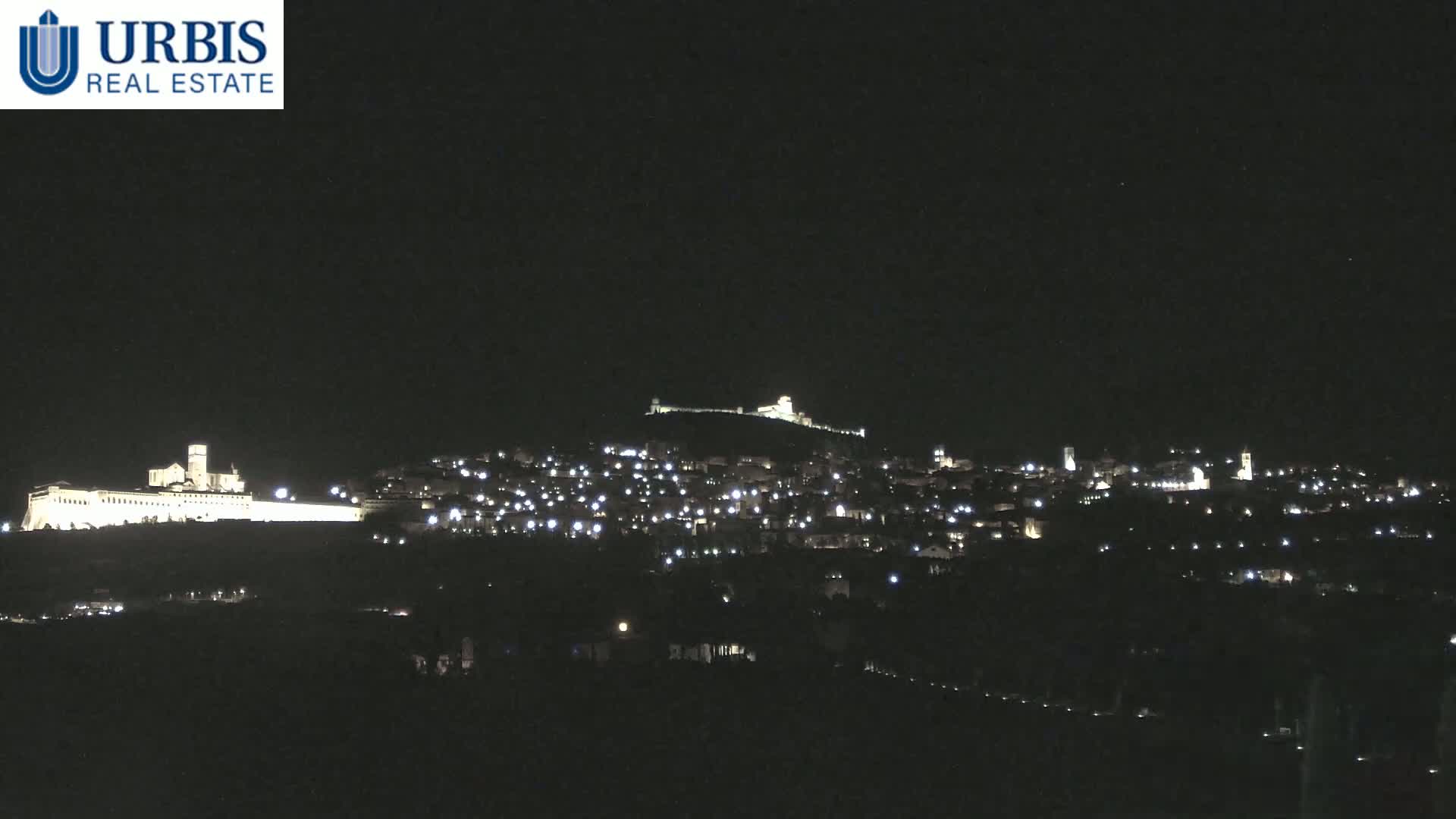 A nighttime view shows a hilltop town illuminated by scattered lights, with a large, brightly lit building on a lower hill in the foreground under a clear, dark sky.