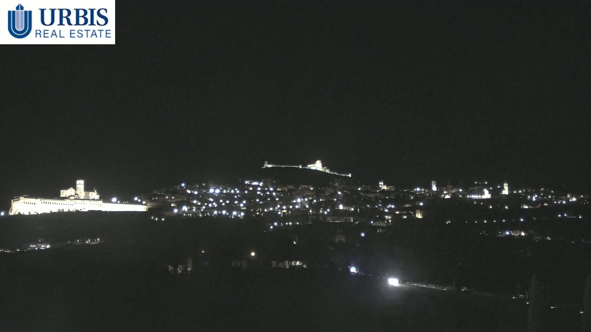 A nighttime long shot shows a hilltop town illuminated with scattered lights, featuring a large, brightly lit building on a lower hill to the left.