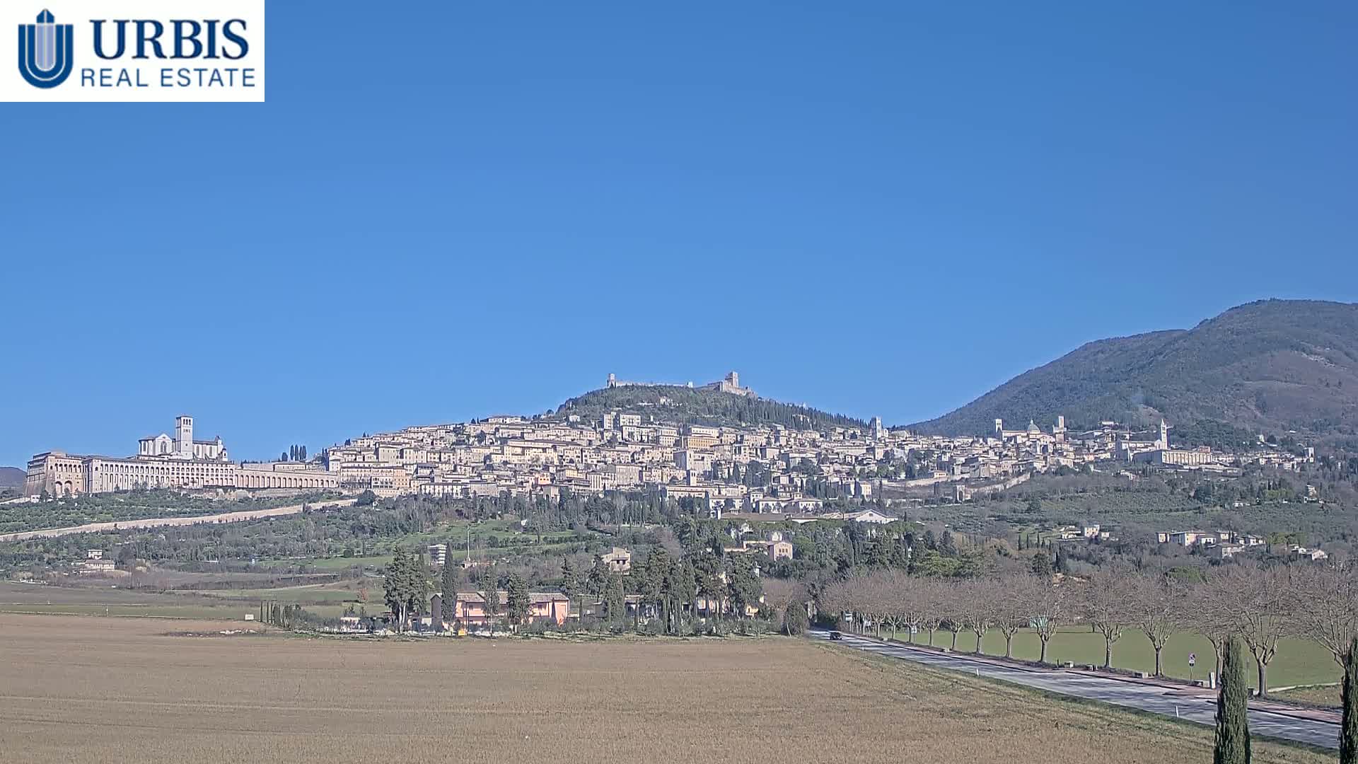 A hilltop town, possibly Assisi, stretches across a hillside under a clear blue sky, with a flat, brown field in the foreground and a mountain in the background.