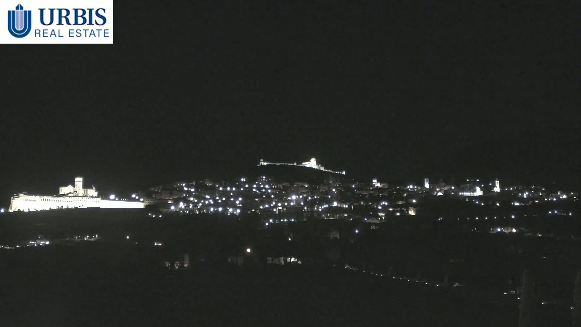A nighttime view shows a hilltop town illuminated with scattered lights, including a large, brightly lit building on the lower left.