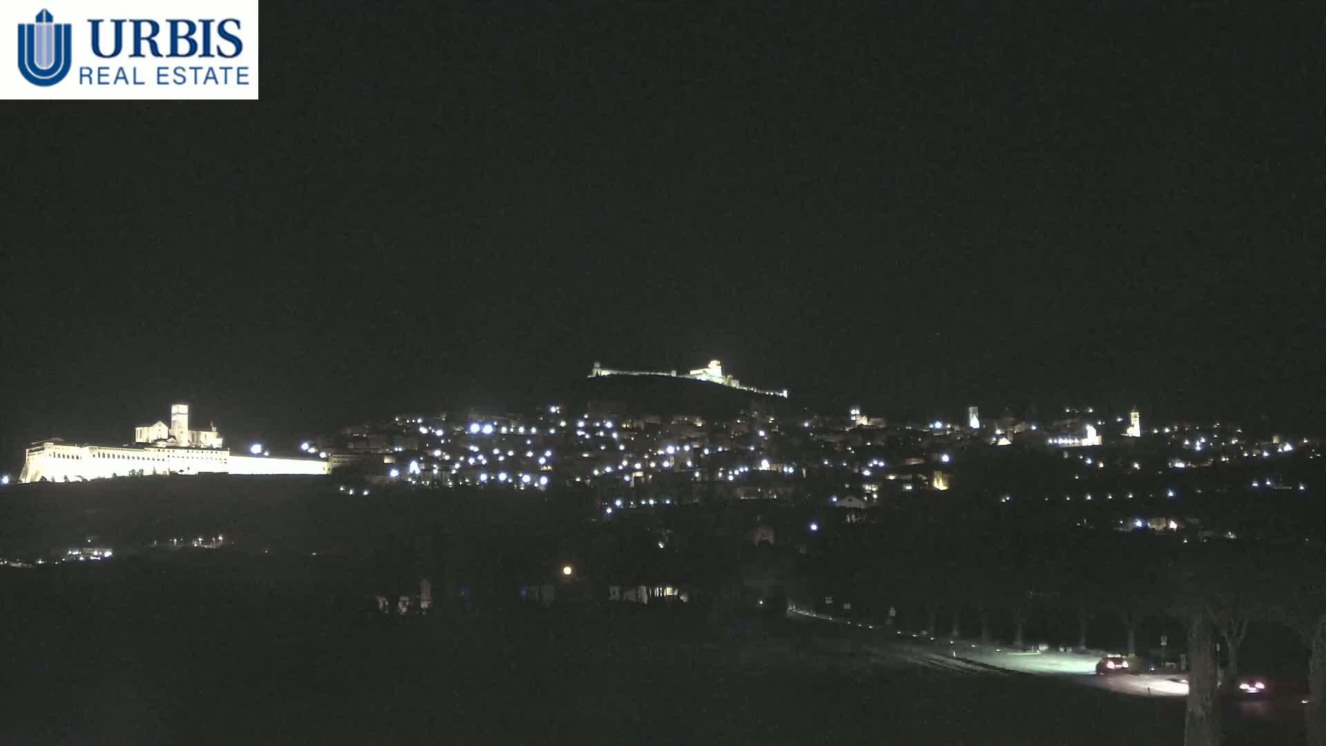 A nighttime view of a hilltop town illuminated by lights, with a large building brightly lit on a lower hill in the foreground under a clear, dark sky.