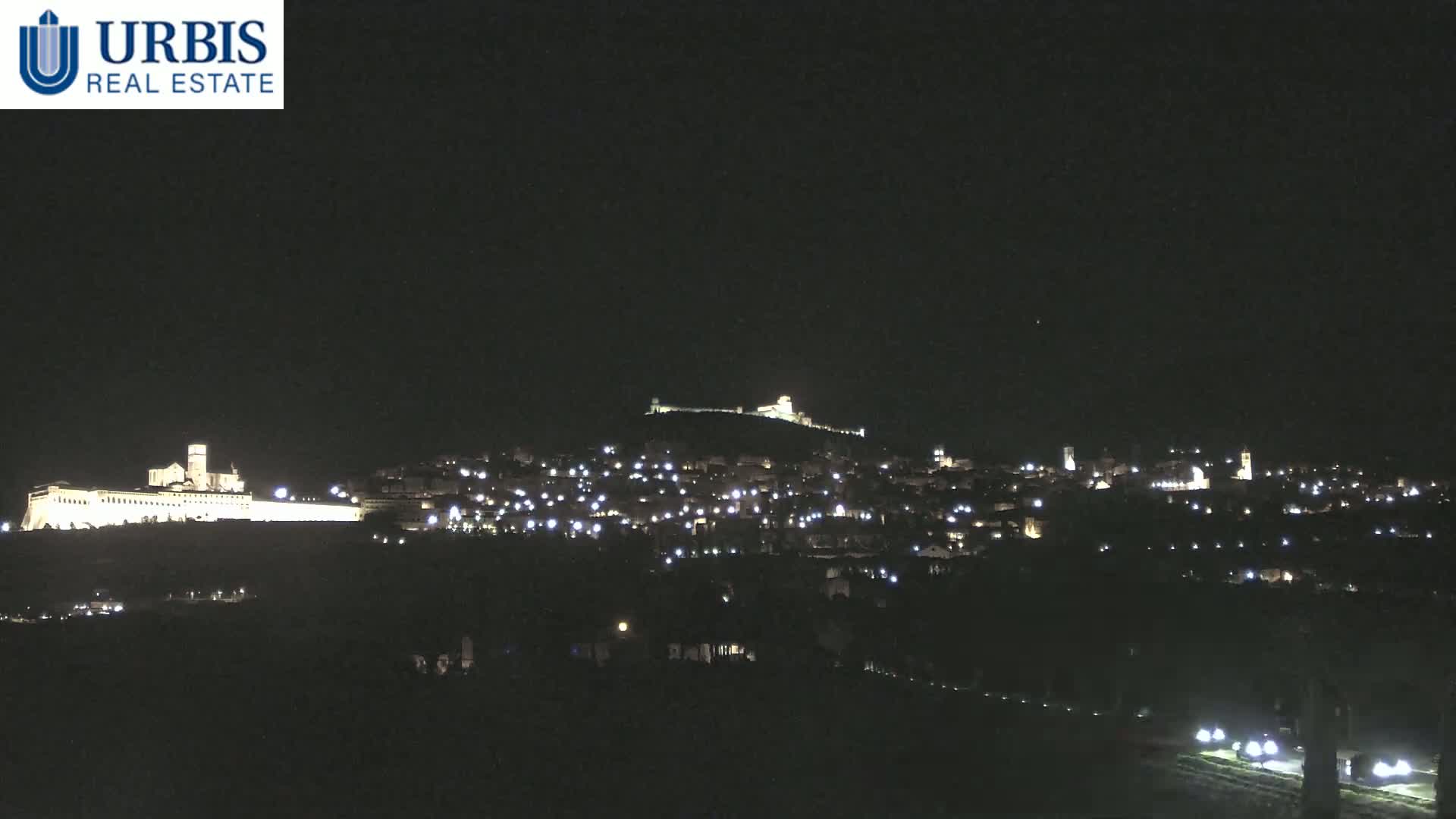 A nighttime view of a hilltop town illuminated by numerous lights, with a large, brightly lit structure at the base of the hill.