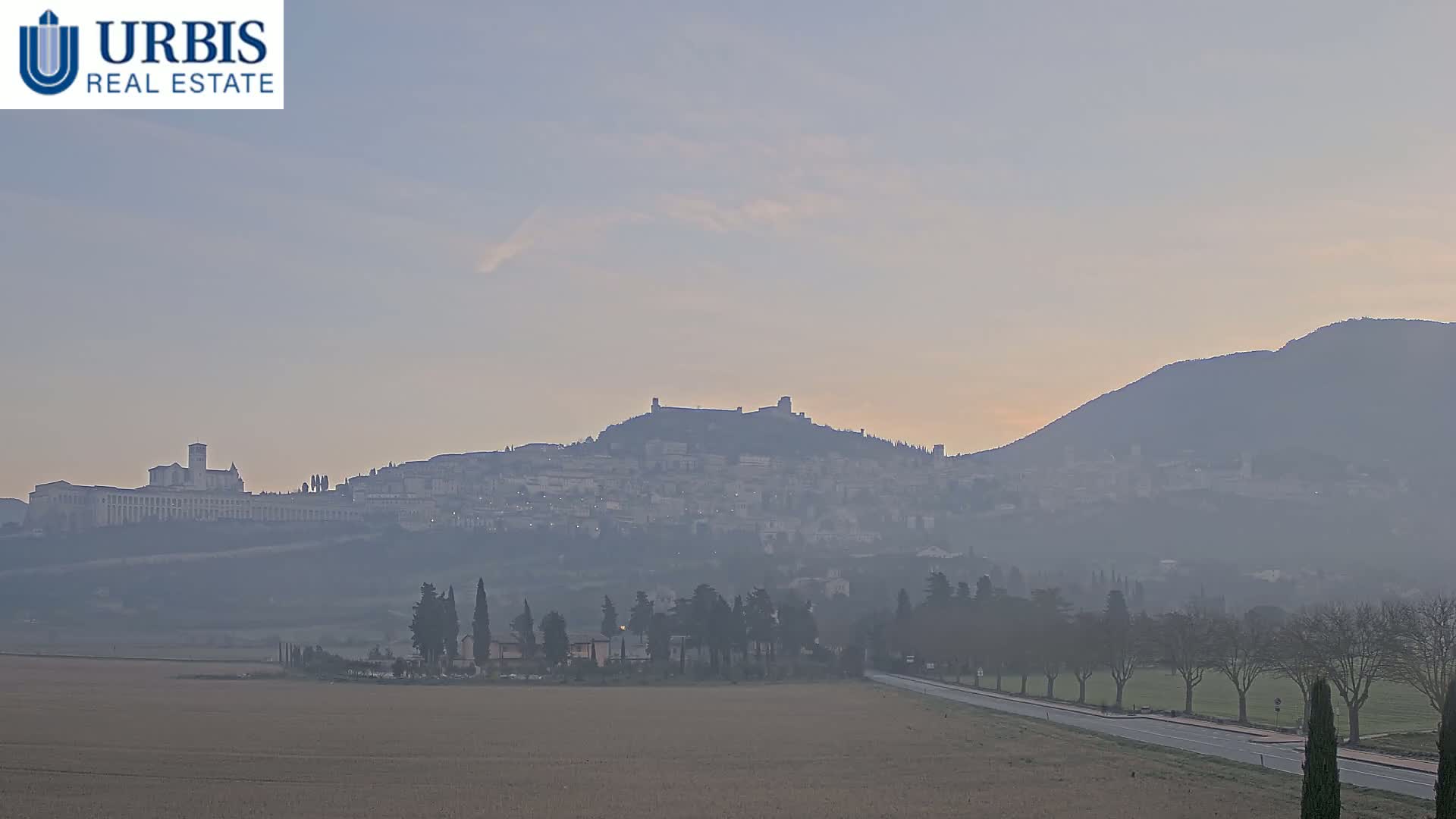 A hazy, pale sunrise illuminates a hilltop town and a large building complex, situated in a valley next to a large mountain.