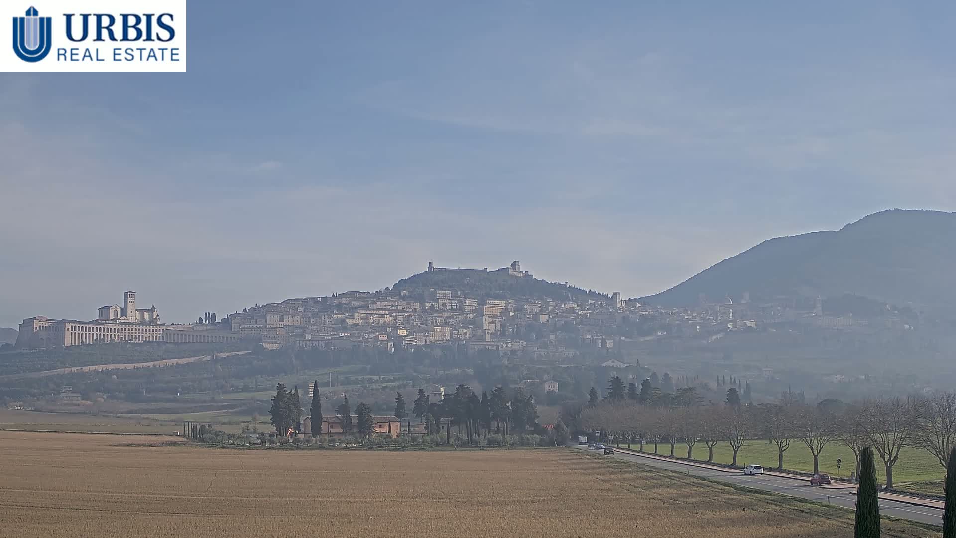 A hazy view of Assisi, Italy, nestled on a hill, with a flat, dry field and road in the foreground under a clear, bright sky.