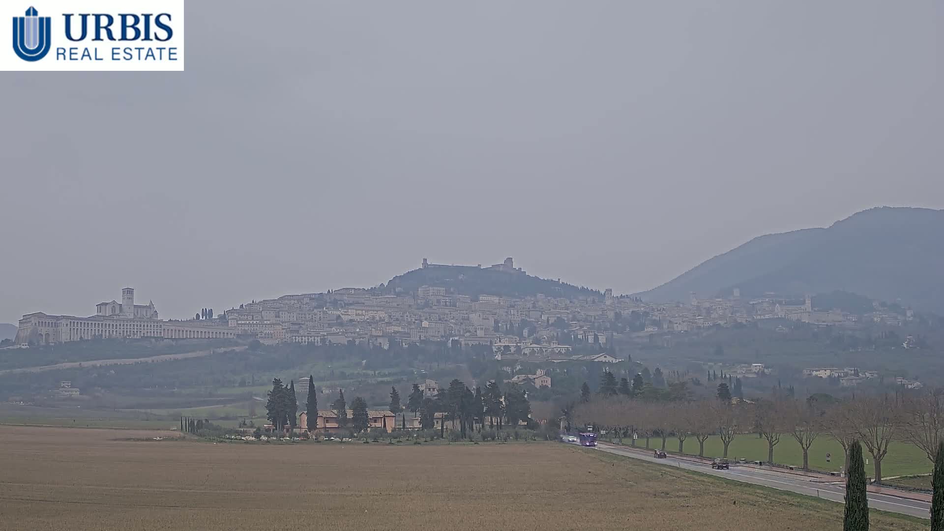 A hazy view of a large town nestled on a hill, with a long, flat building at its base, in the distance, and a flat field in the foreground with a road running through it.