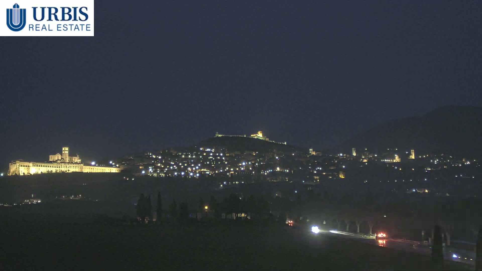 A nighttime view of a hilltop town illuminated by lights, with a large building complex also lit up on a lower hill in the distance, under a dark, clear sky.