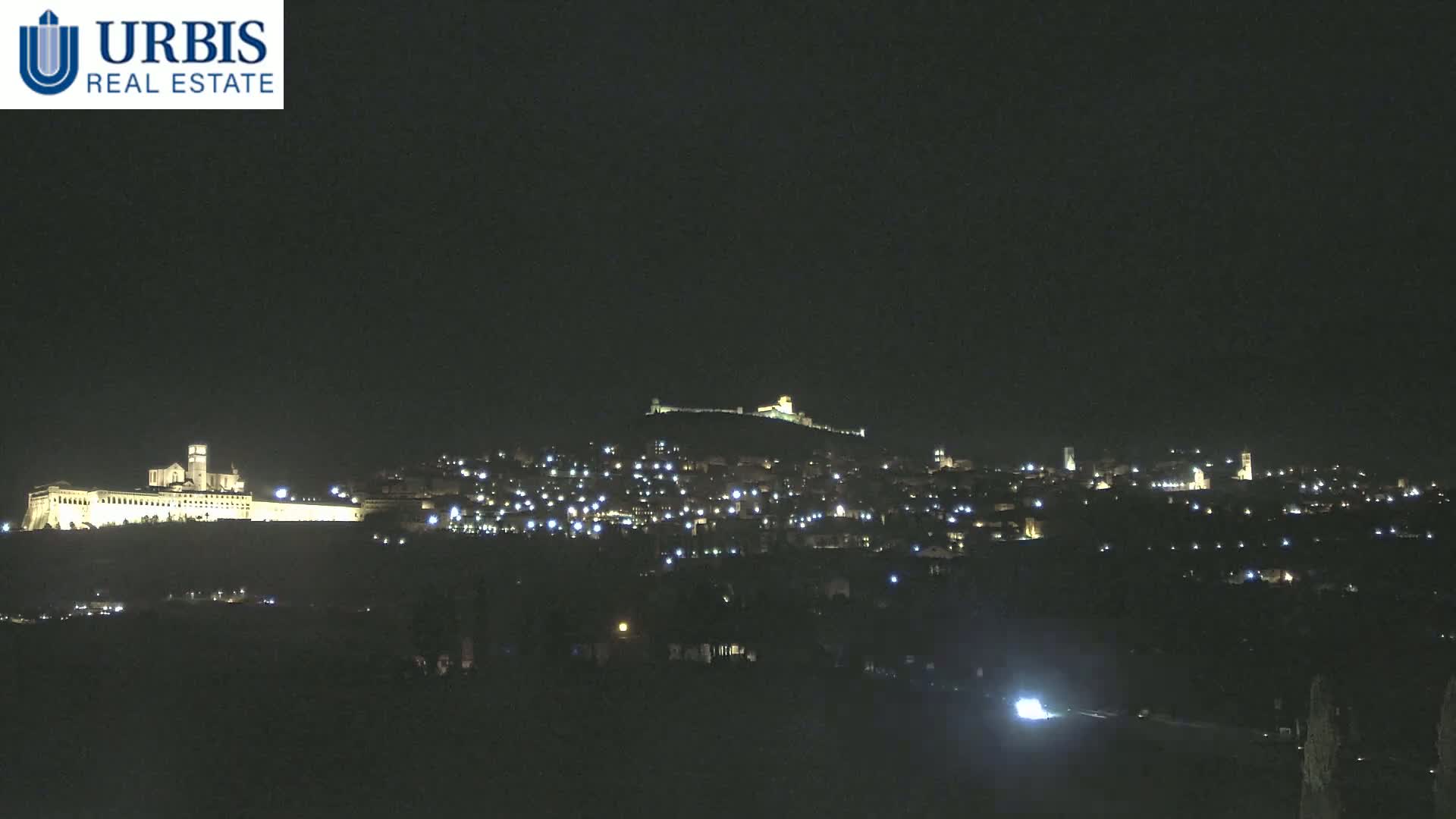A nighttime view of a hilltop town illuminated by scattered lights, featuring a large, brightly lit building complex at the base of the hill.